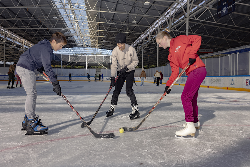 Drie kinderen spelen met ijshockeysticks tijdens het recreatief schaatsen bij een Optisport Schaatsbaan