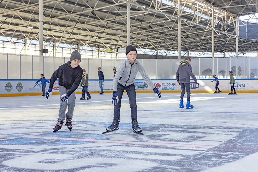 Twee jongetjes schaatsen lachend richting de camera tijdens het recreatief schaatsen bij een Optisport Schaatsbaan