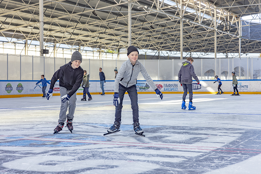 Twee jongetjes schaatsen lachend richting de camera tijdens het recreatief schaatsen bij een Optisport schaatsbaan