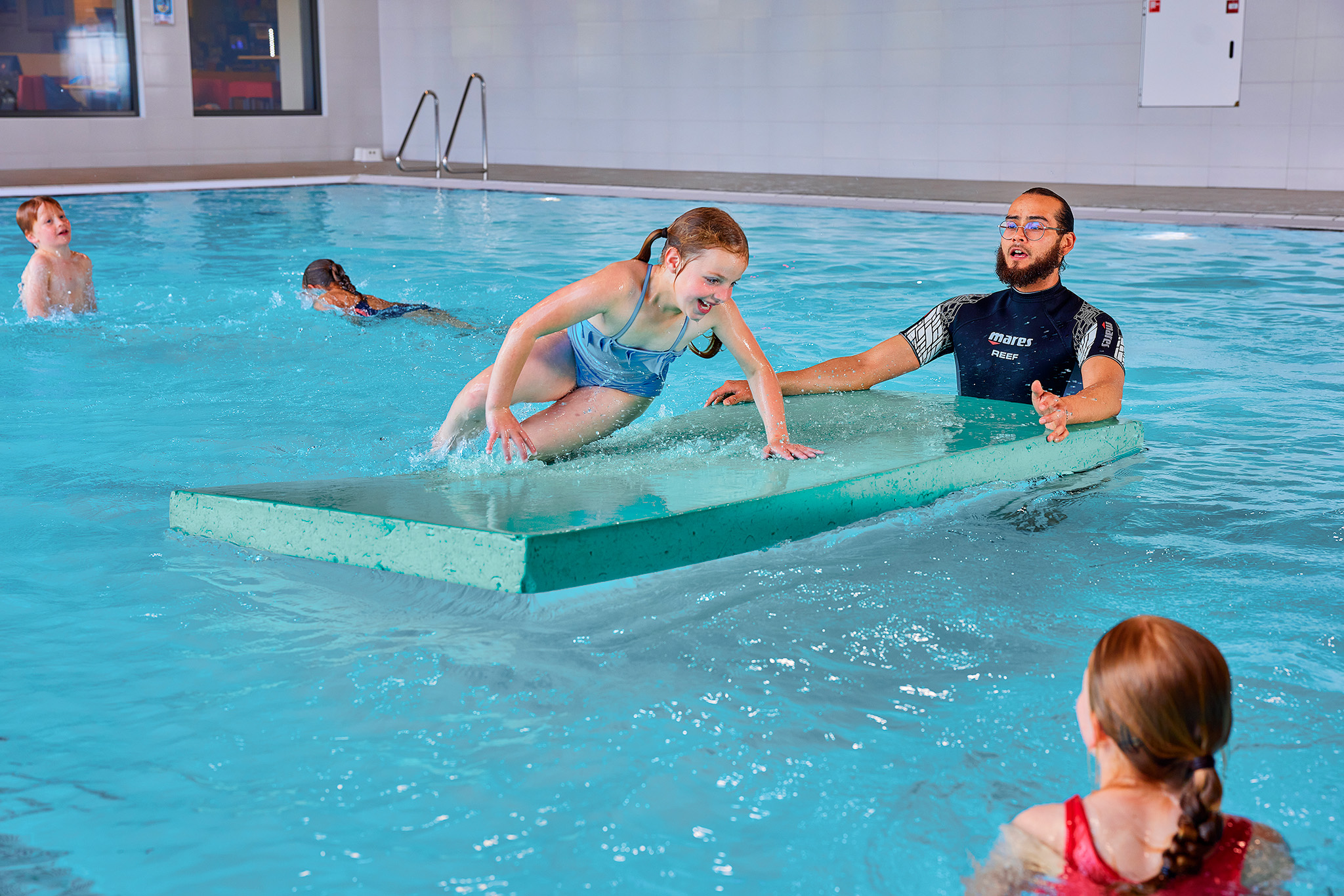 Meisje zit op een mat in het water met een man erlangs die in het water staat tijdens Aqua'Fun