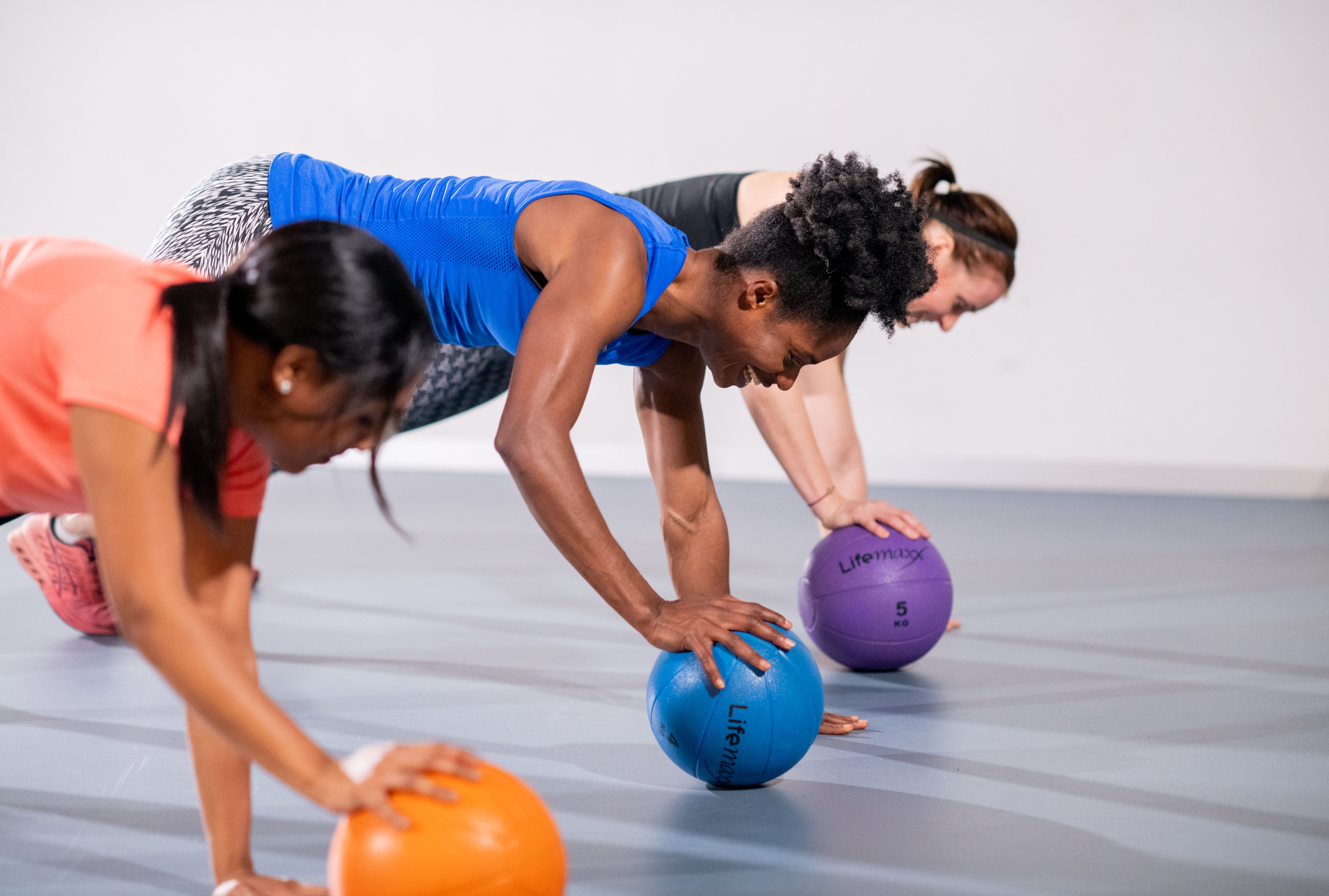 Drie vrouwen doen oefeningen met een bal en hebben plezier tijdens het trainen bij een Optisport locatie