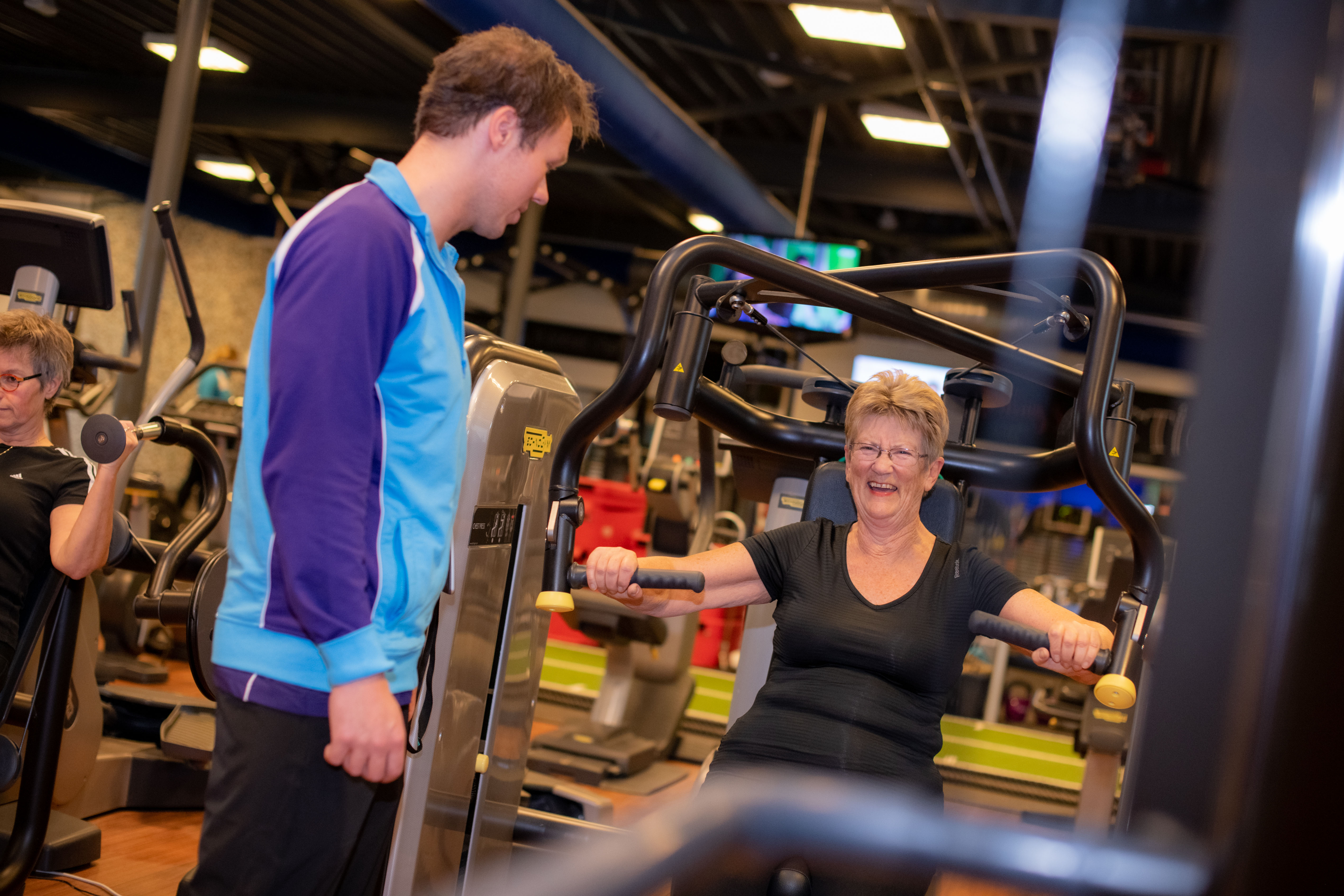 Fitness bij Optisport. Seniorenfitness, een vrouw is oefeningen aan het doen onder begeleiding van een instructeur.