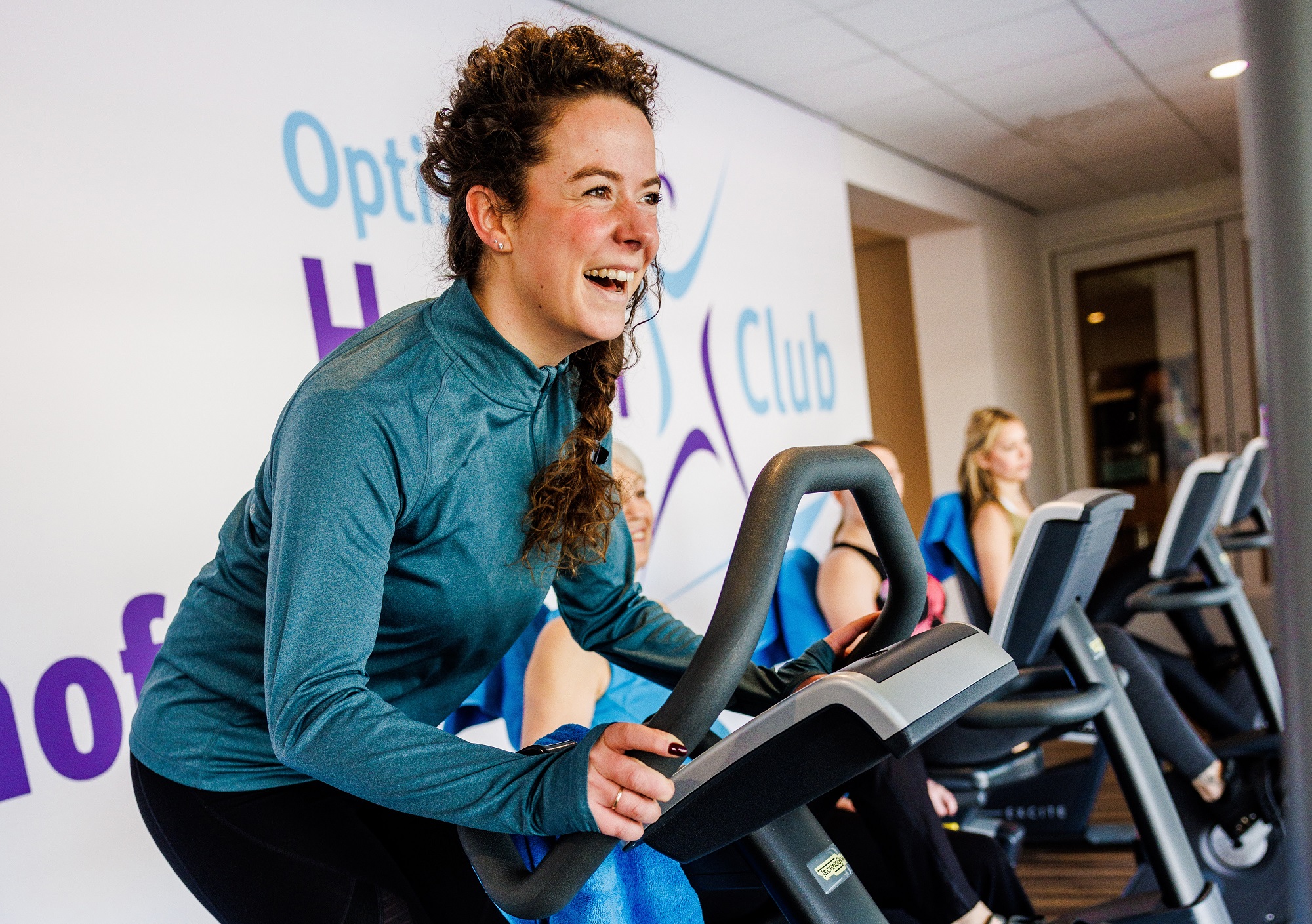 Een jonge sportende dame zit met een enorme glimlach op de hometrainer en geniet van een sportief fietsritje bij Optisport in de Health Club.