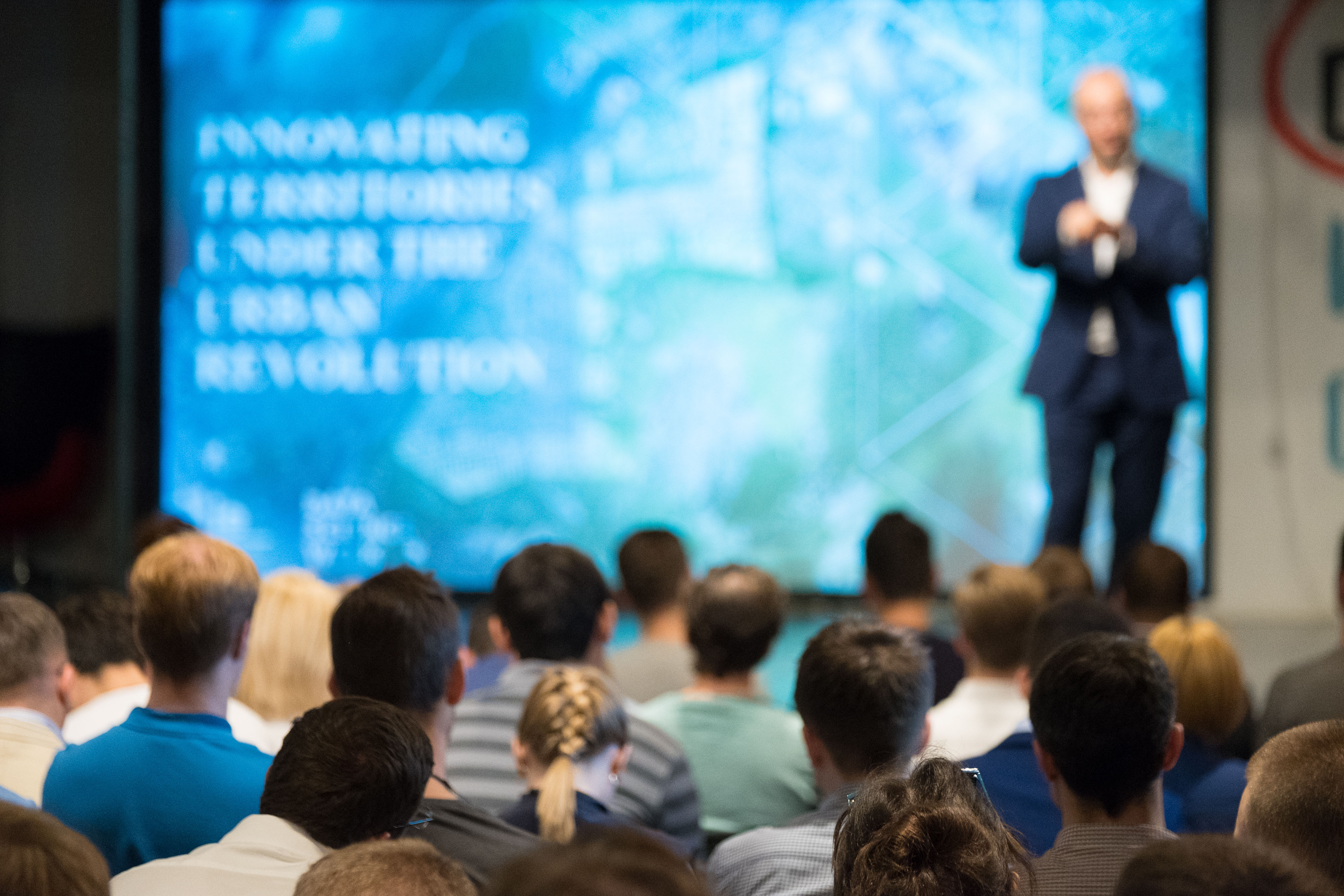 Man op podium geeft presentatie aan groep in de zaal tijdens georganiseerd evenement.