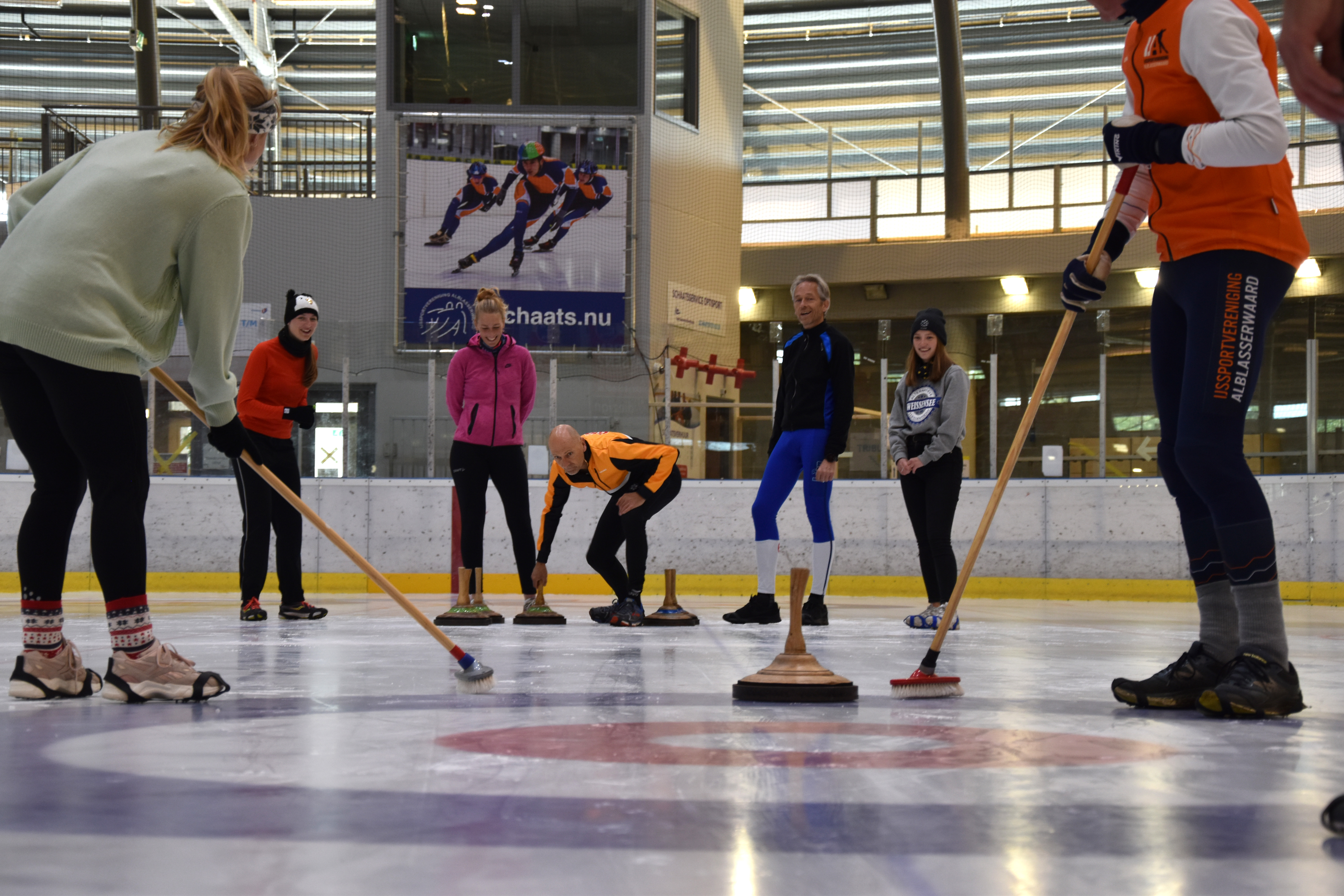 Zeven mensen op het ijs zijn bezig met een potje curling tijdens een schaats uitje 