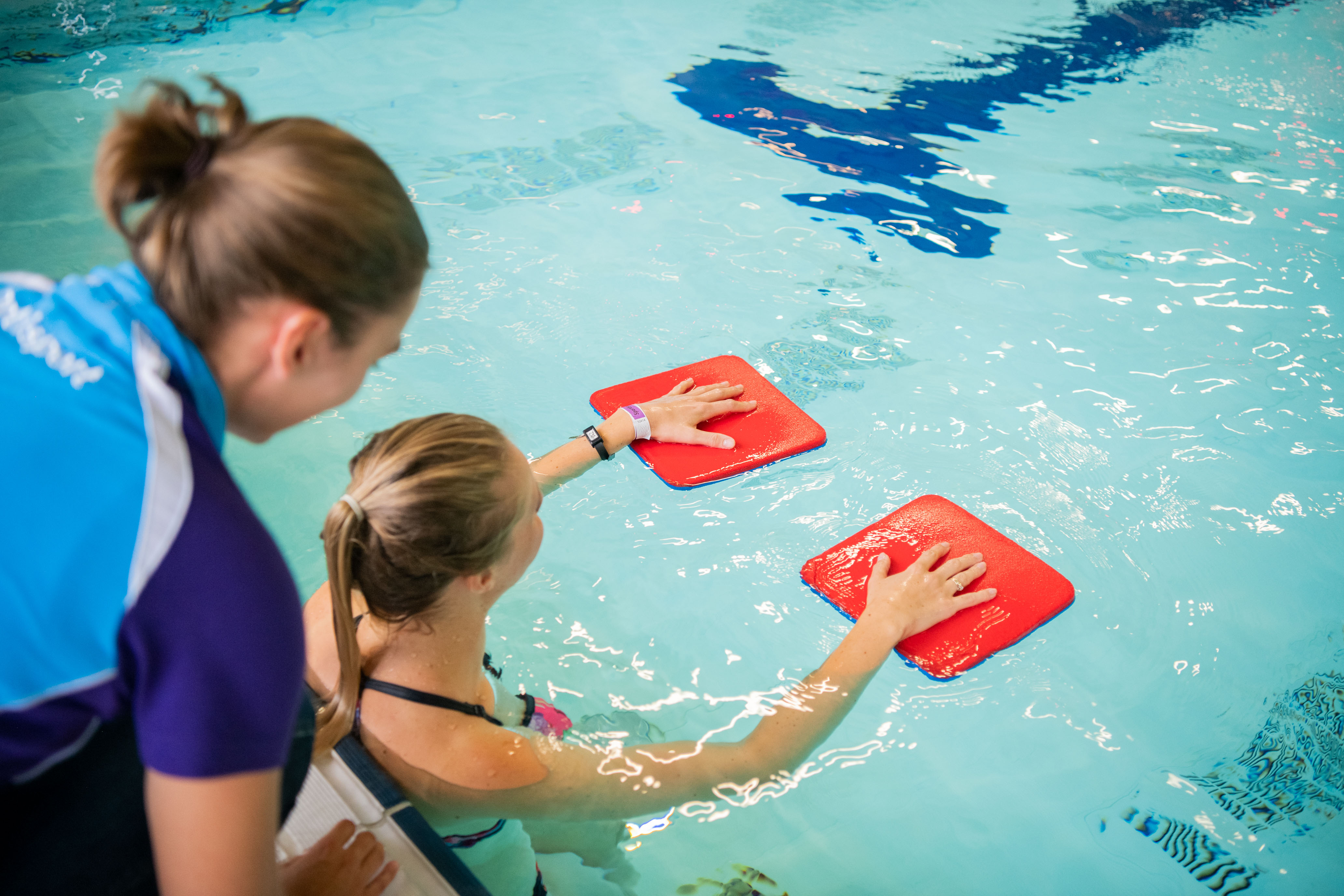 Vrouw heeft haar handen op twee plankjes in het water. Achter haar staat een instructrice