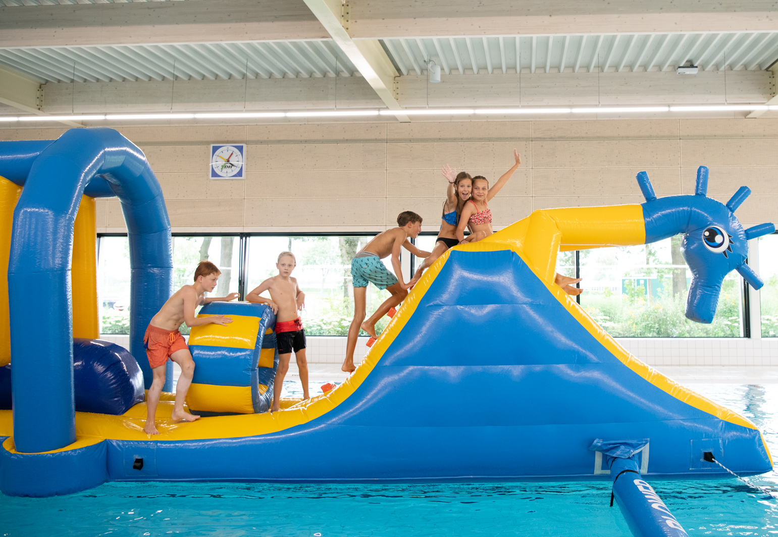 Kinderen hebben plezier in het zwembad van Optisport met de waterspeeltuin. Ze klimmen op een opblaaskussen onder toezicht van een instructeur.