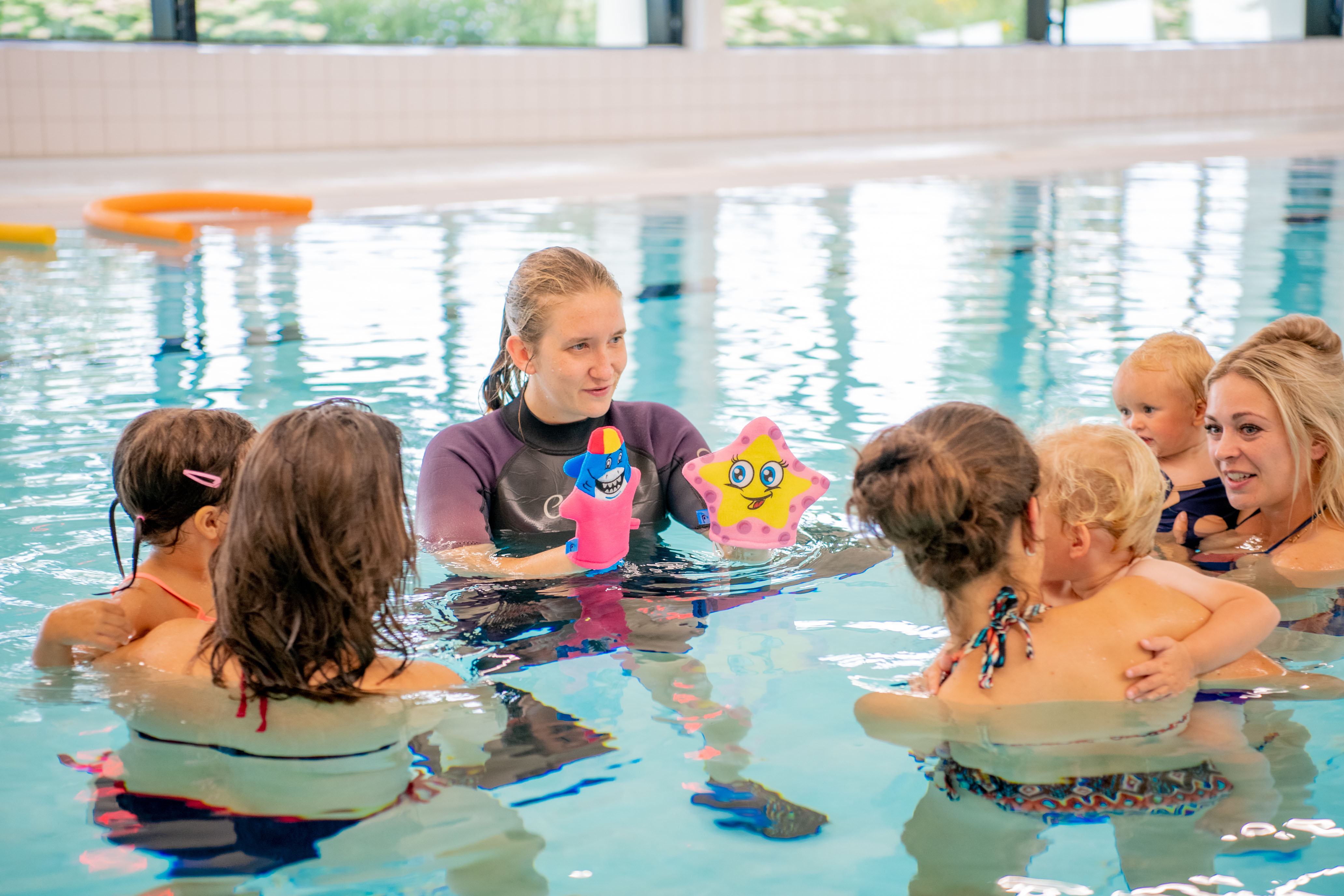 Een groep kinderen zitten vrolijk in het water met hun ouders bezig terwijl de instructeur van Optisport hierbij is.