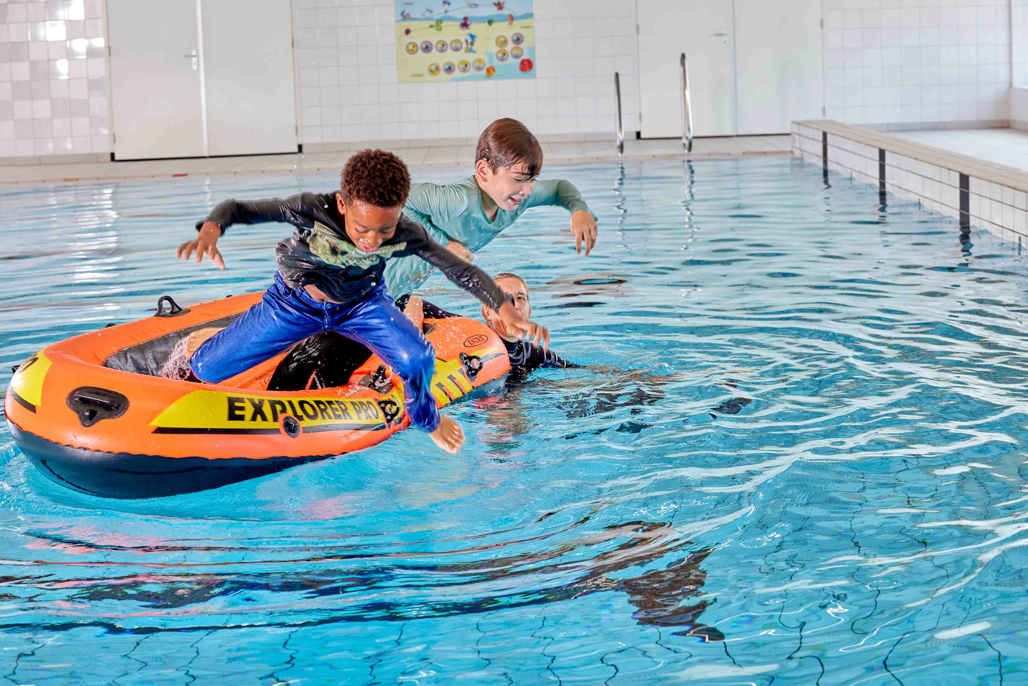Twee jongetjes springen, geheel aangekleed, uit een bootje het zwembad van Optisport in terwijl de instructeur toekijkt. 