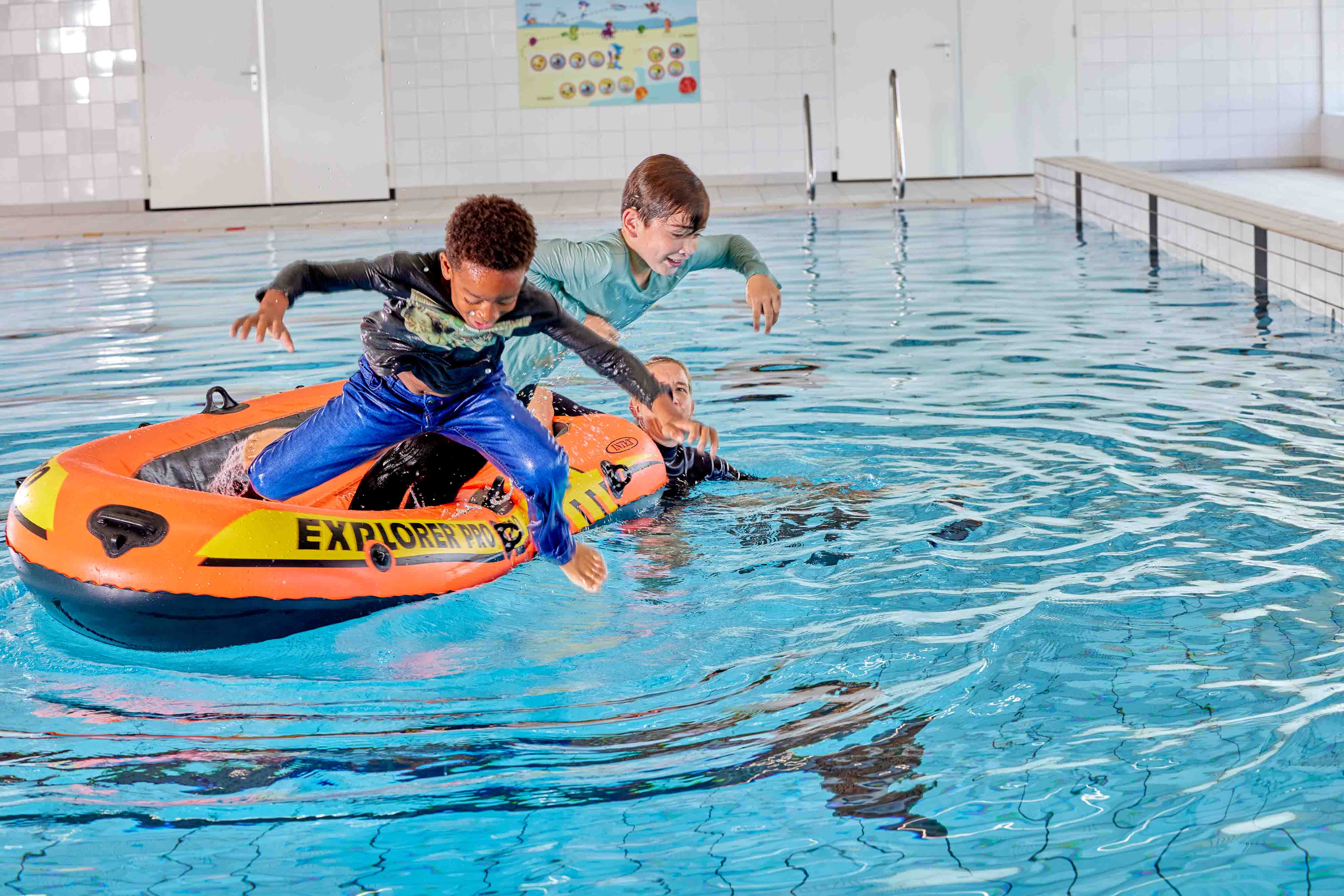 Twee jongetjes springen, geheel aangekleed, uit een bootje het zwembad van Optisport in terwijl de instructeur toekijkt.