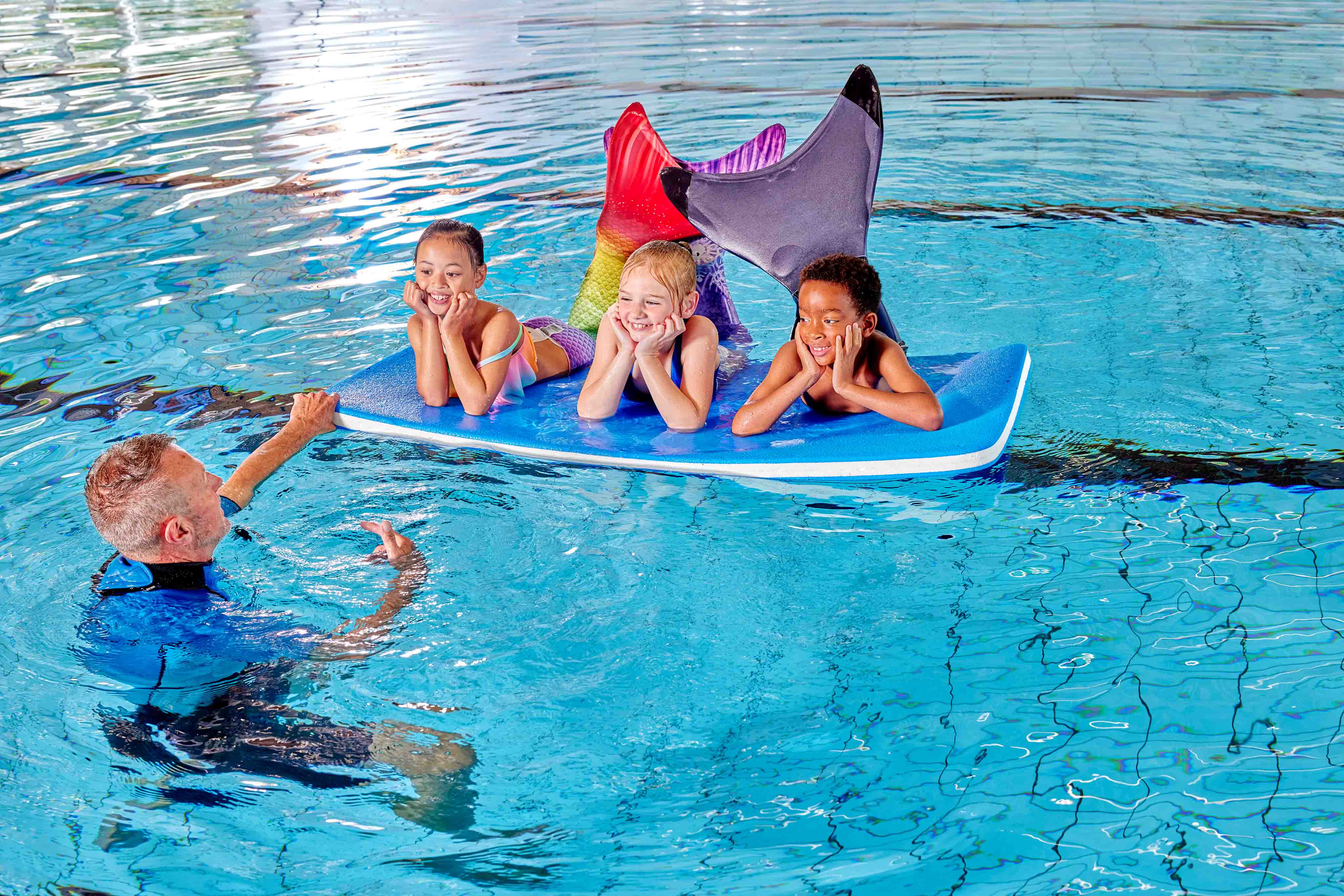 Drie meiden en een jongen met zeemeerminstaarten leunen met hun ellebogen op de rand van het zwembad van Optisport, tijdens de les Zeemeerminzwemmen. 