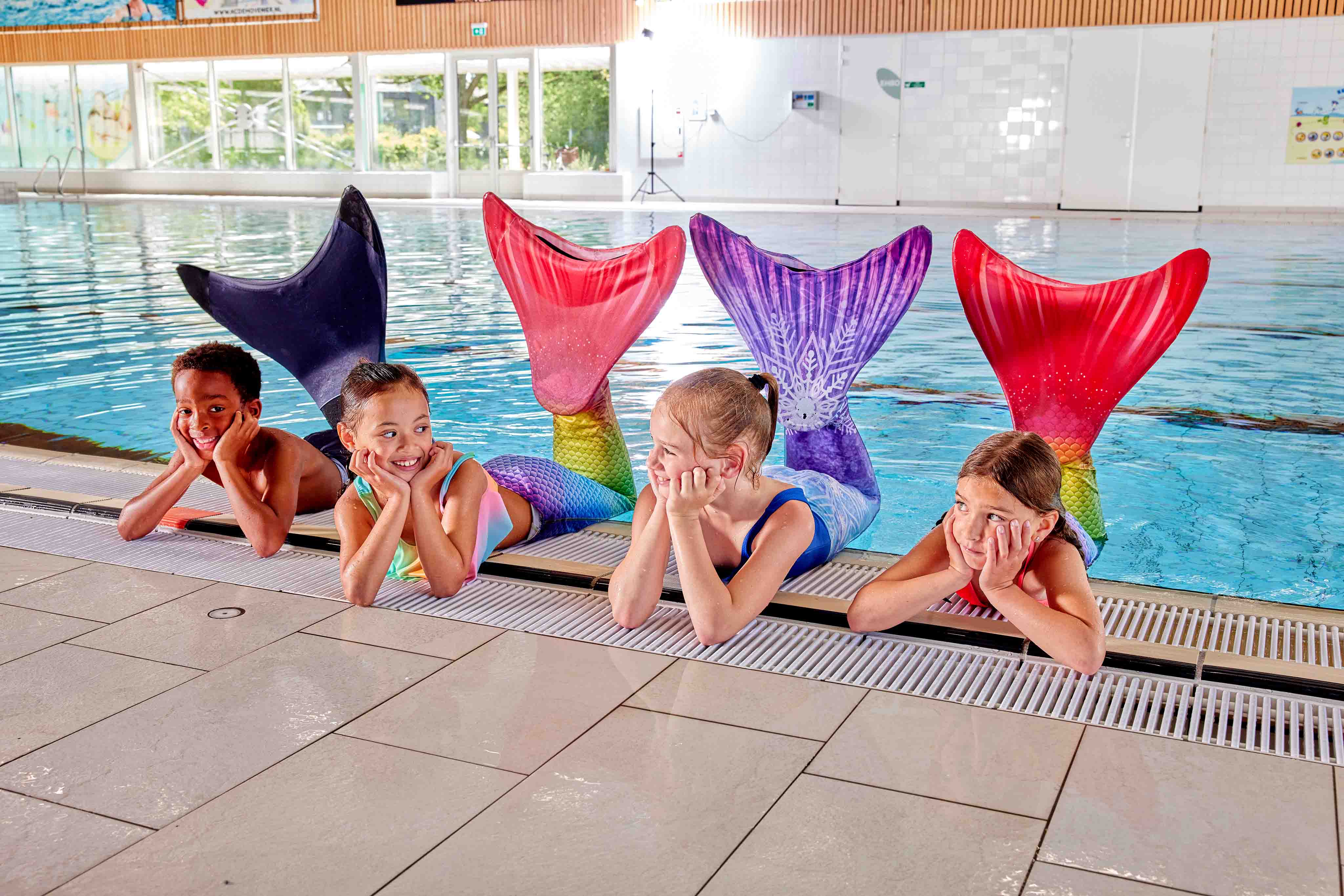 Vier kinderen zitten aan de rand van een Optisport zwembad te poseren met een zeemeerminstaart.