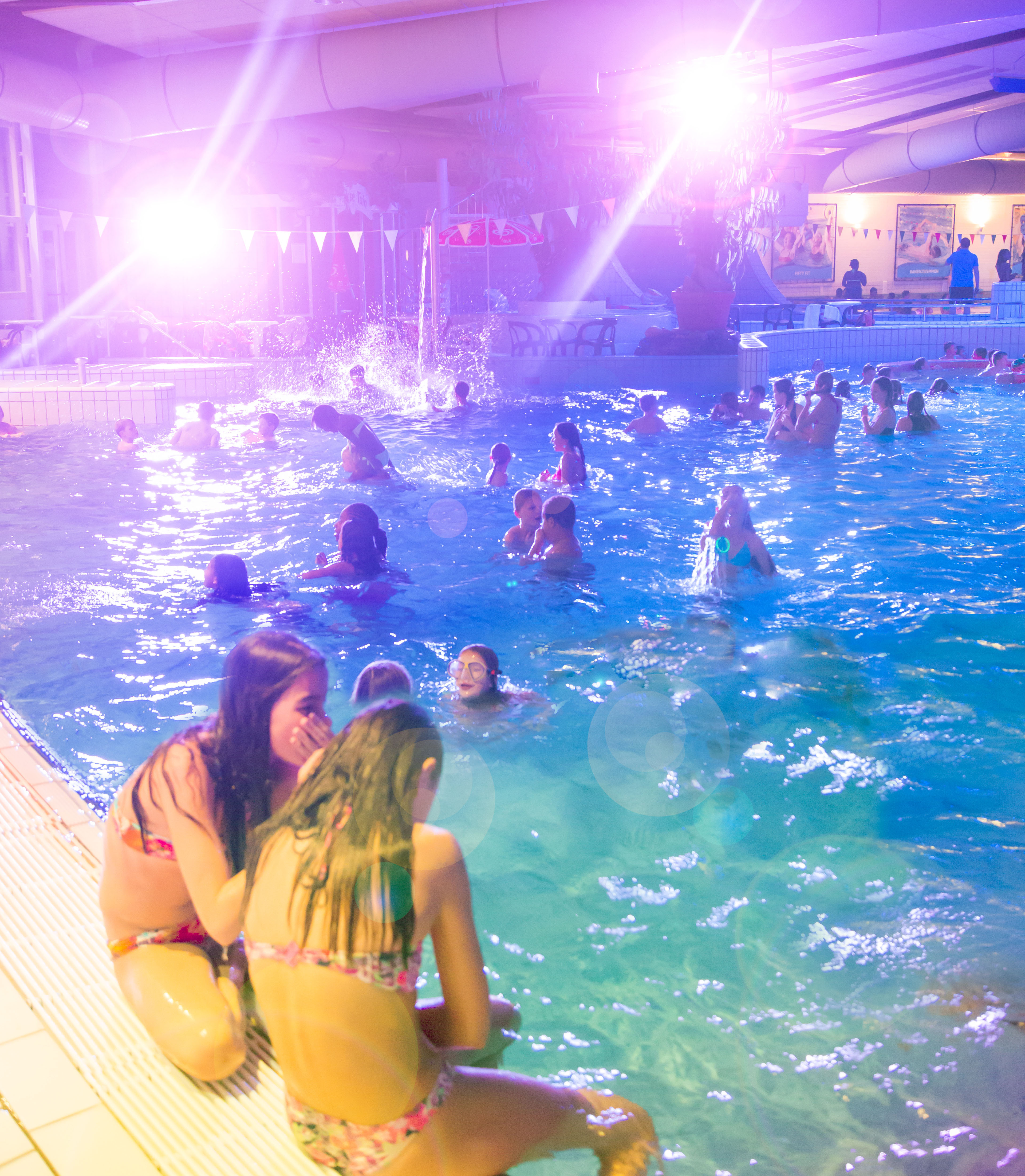 Verschillende kinderen zwemmen in het water tijdens een zwemdisco bij Optisport. Vooraan in de foto zitten twee meisjes op de rand.