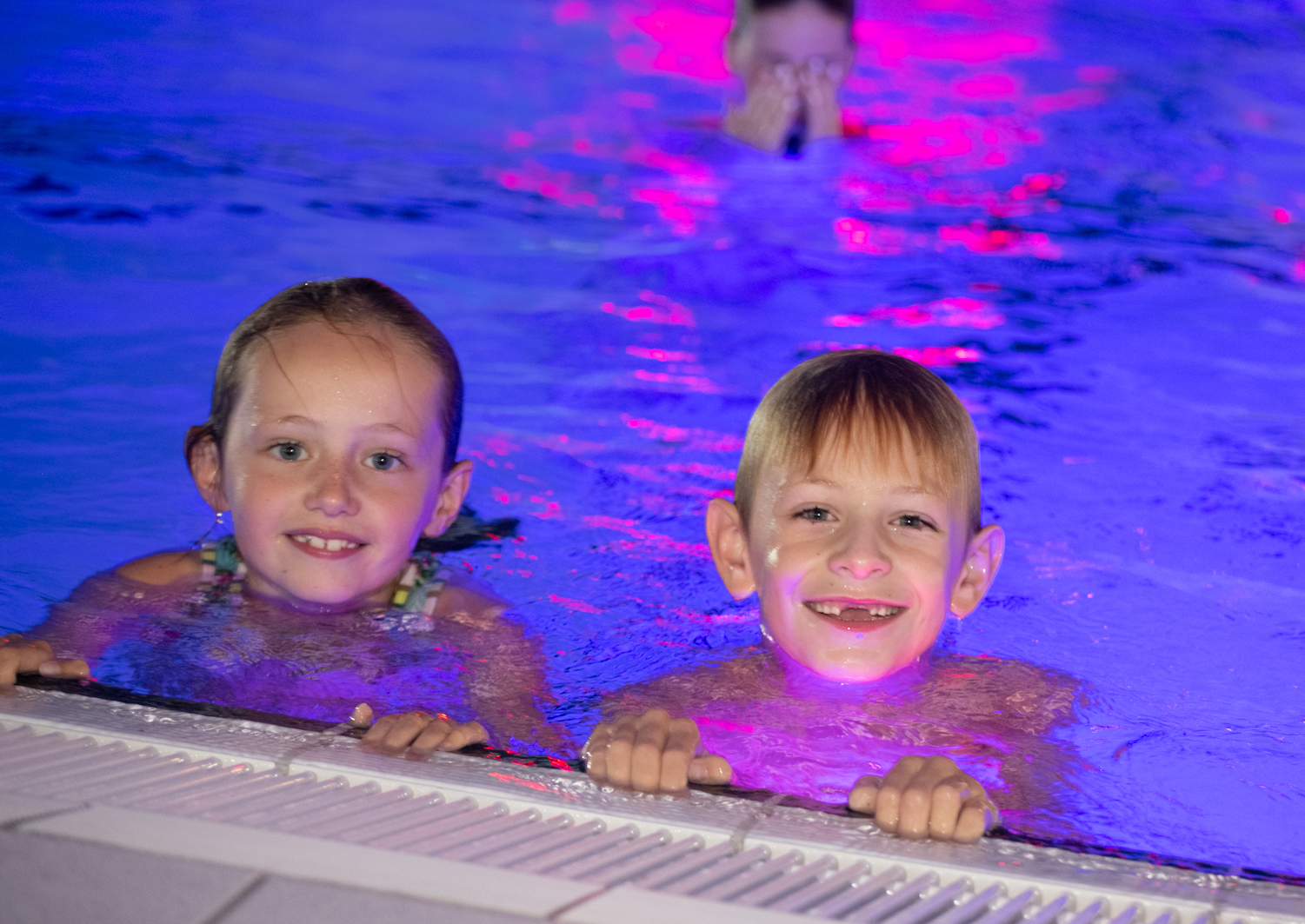 Twee kinderen hangen aan de rand en kijken blij in de camera tijdens het zwemdiscofeestje.