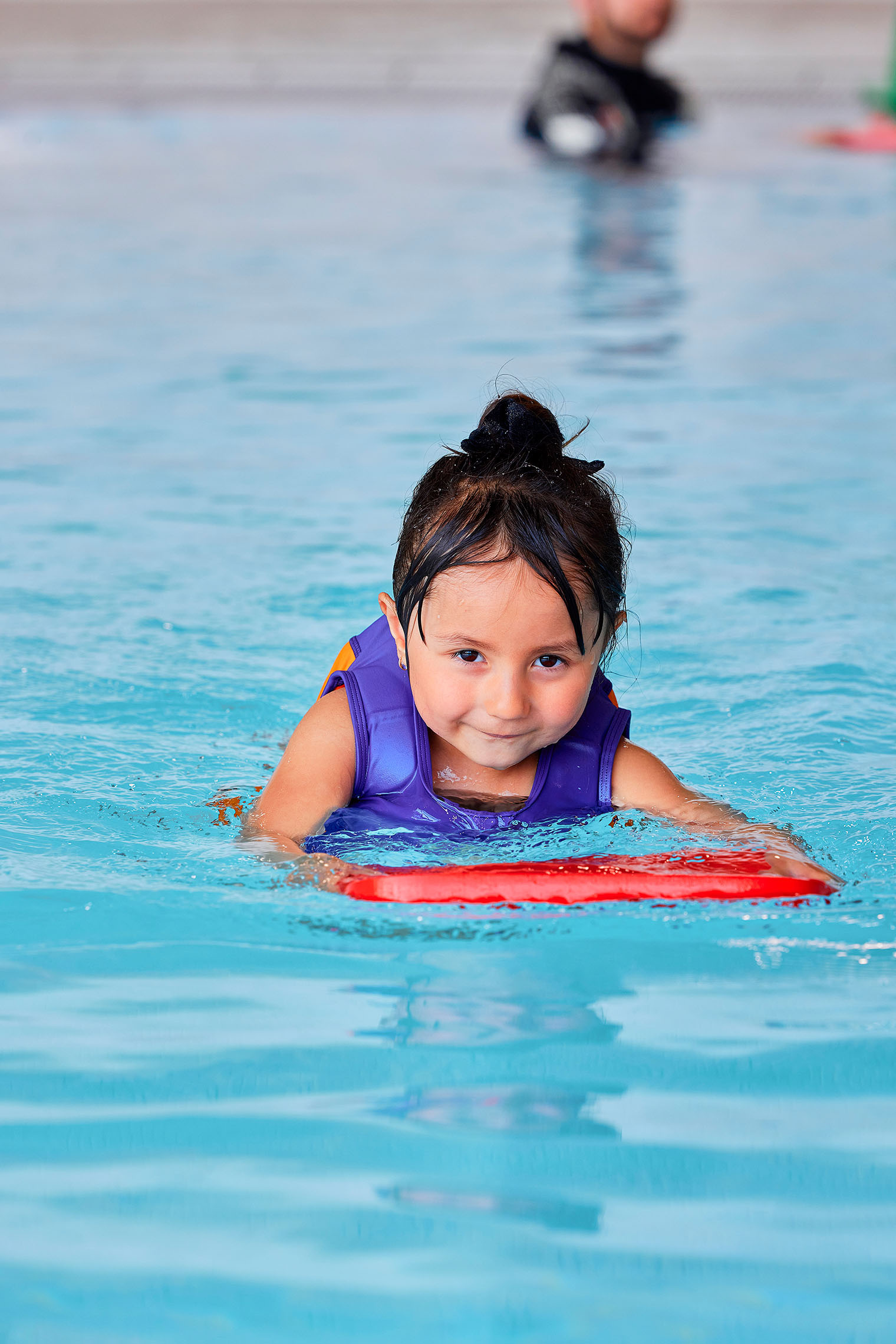 Een klein meisje kijkt trots in de camera terwijl ze met haar plankje zwemt tijdens een Zed & Sop zwemles bij Optisport. 