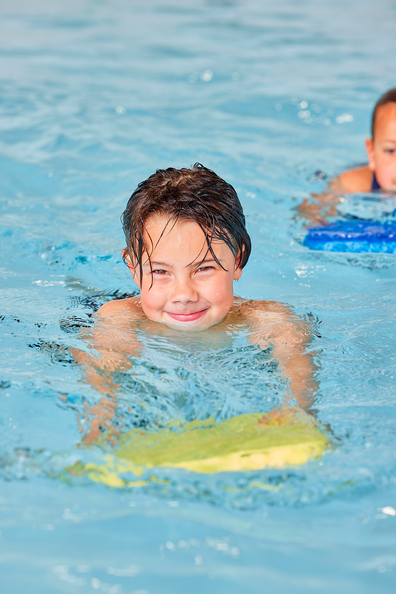 Jongentje zwemt in het water met een drijfplankje en kijkt lachend in de camera.