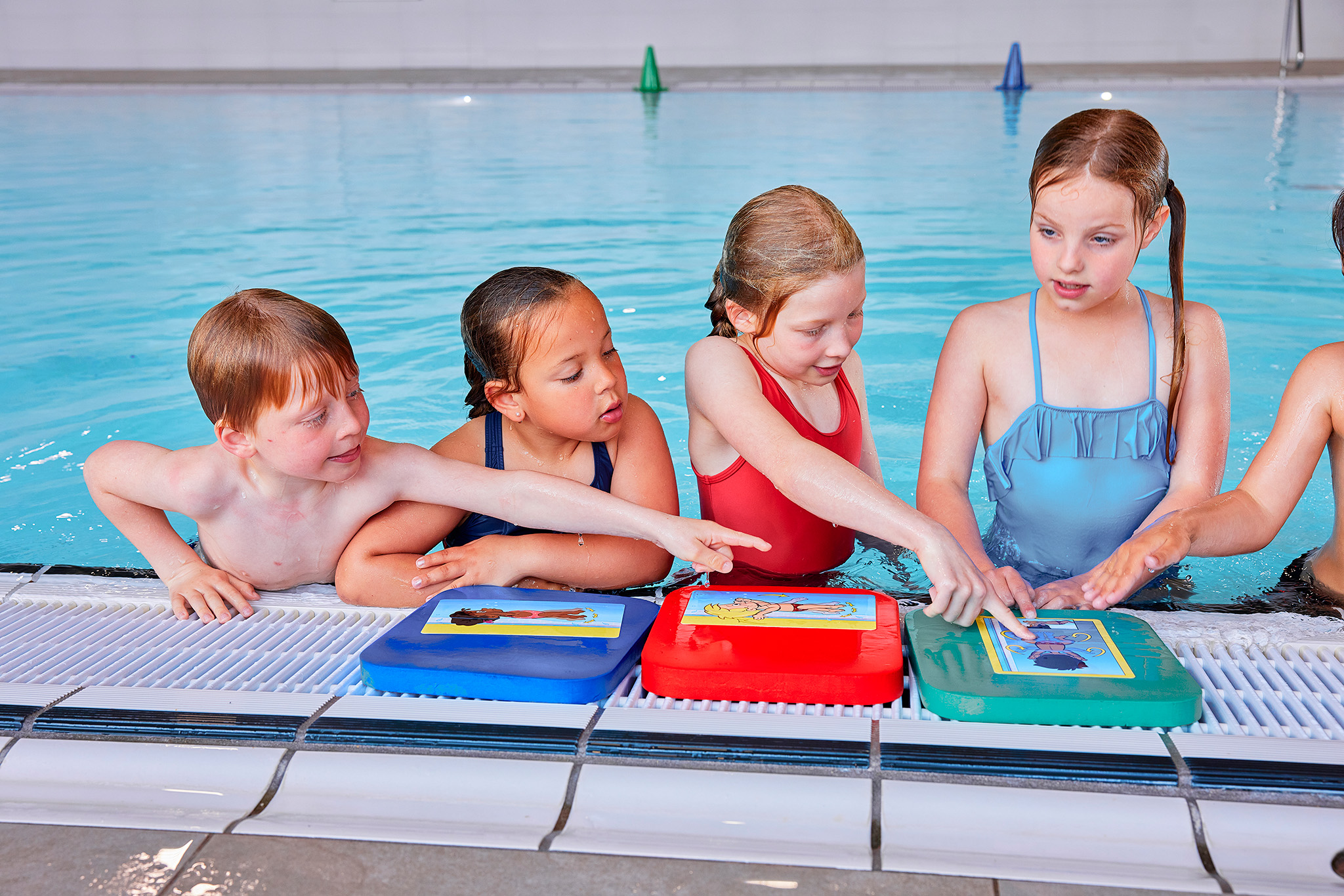 Kinderen bespreken zwemkaarten met instructies langs de rand van het zwembad tijdens de Zed & Sop zwemles bij Optisport. 