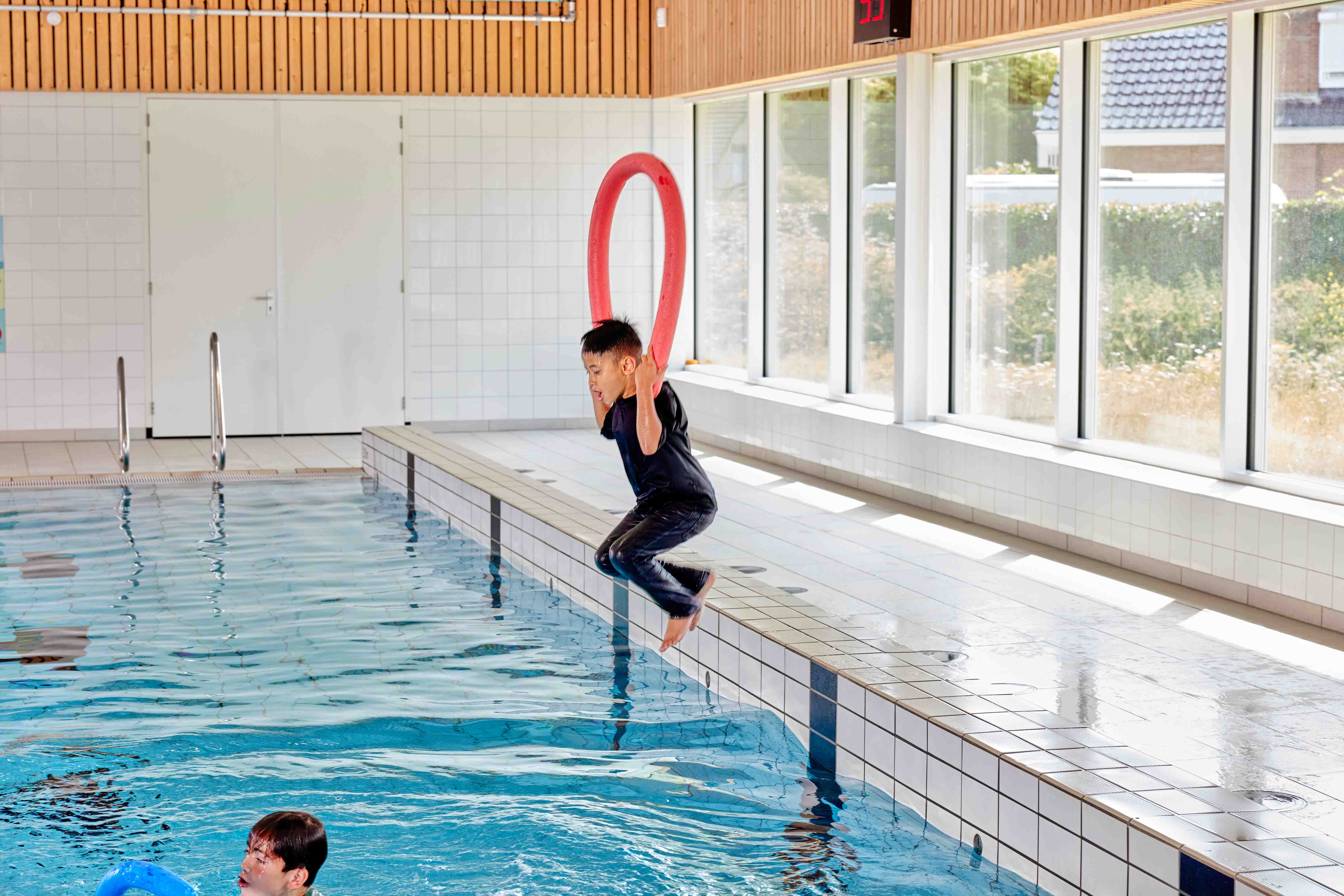 Geheel gekleed springt een jongetje het zwembad in terwijl hij een drijfmiddel vasthoudt, tijdens de Survival zwemles bij Optisport. 