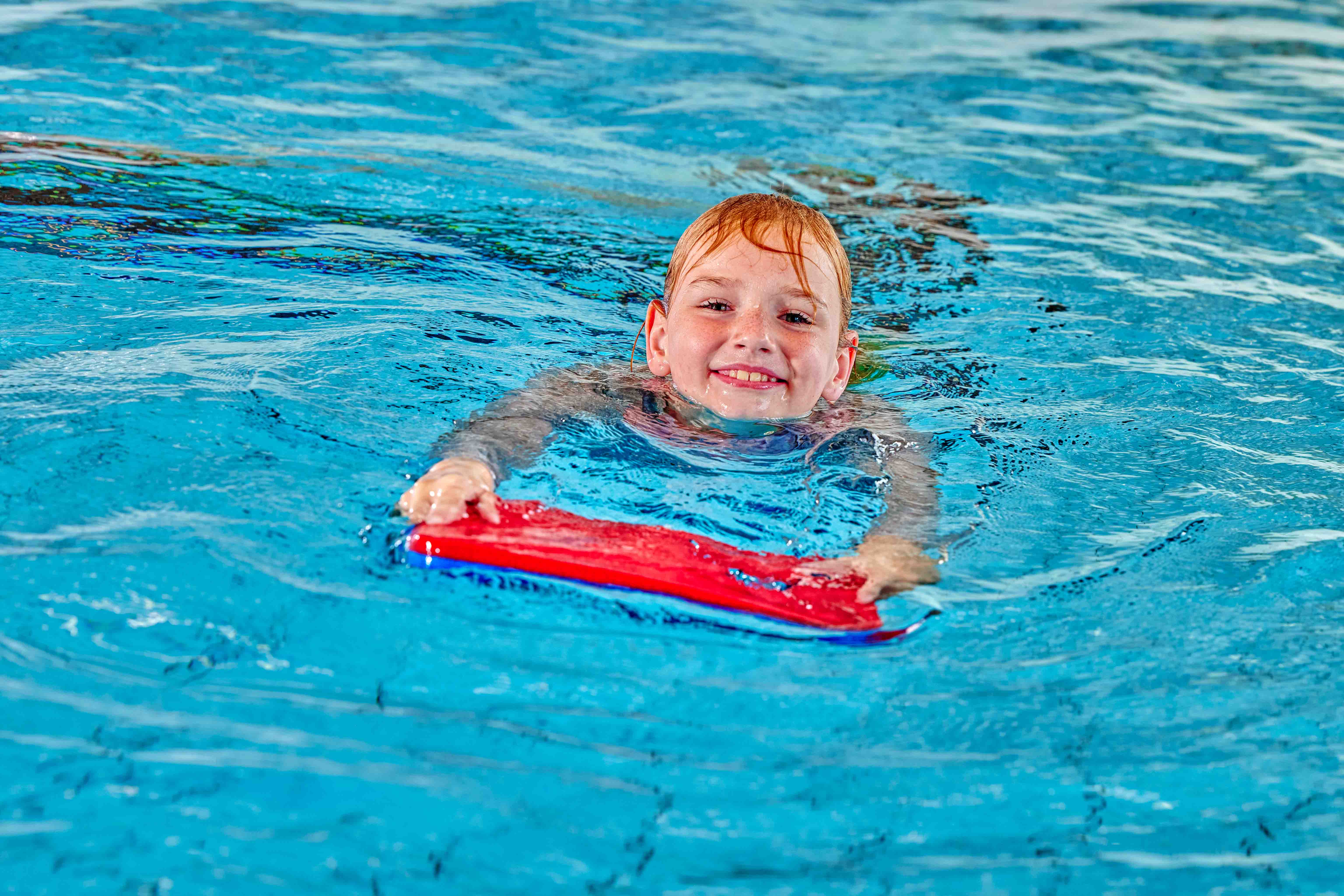 Een jongetje lacht in de camera terwijl hij met zijn zwemplankje zwemt tijdens de les Survival Zwemvaardigheid bij Optisport. 