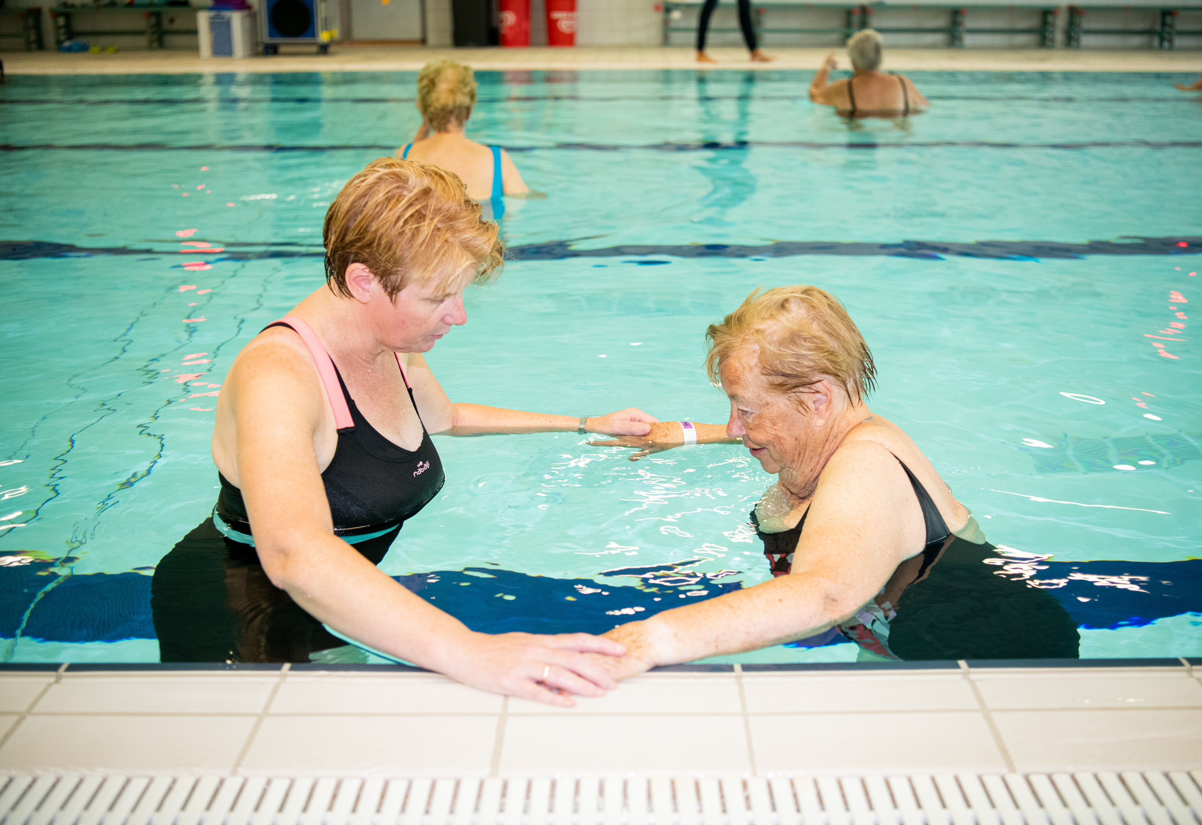 Twee vrouwen hand in hand in het water aan de zijkant van het zwembad doen oefeningen tijdens Aqua'Therapie zwemmen voor hartpatiënten.