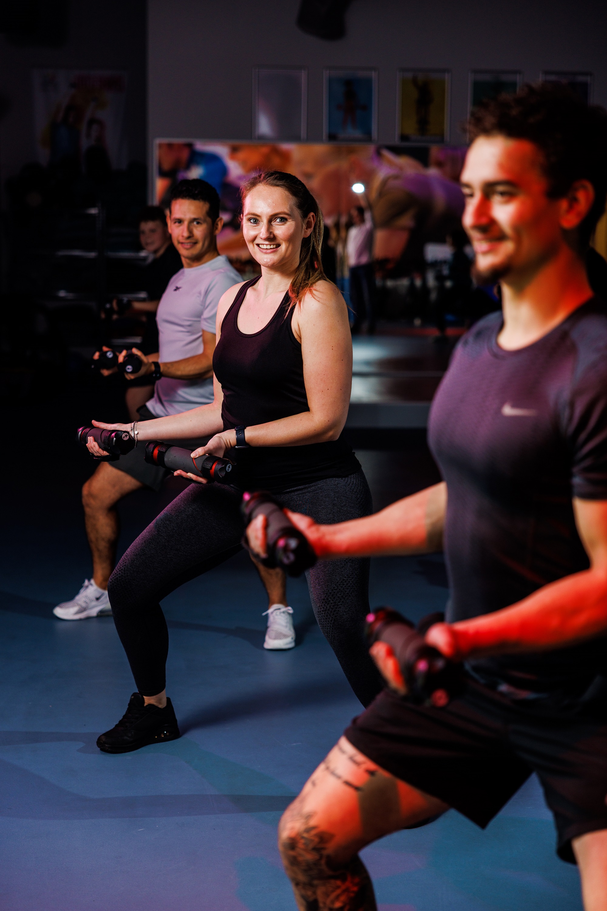 Drie sporters doen intensieve workouts tijdens een groepsles, terwijl ze halters vasthouden en zichtbaar plezier hebben. 