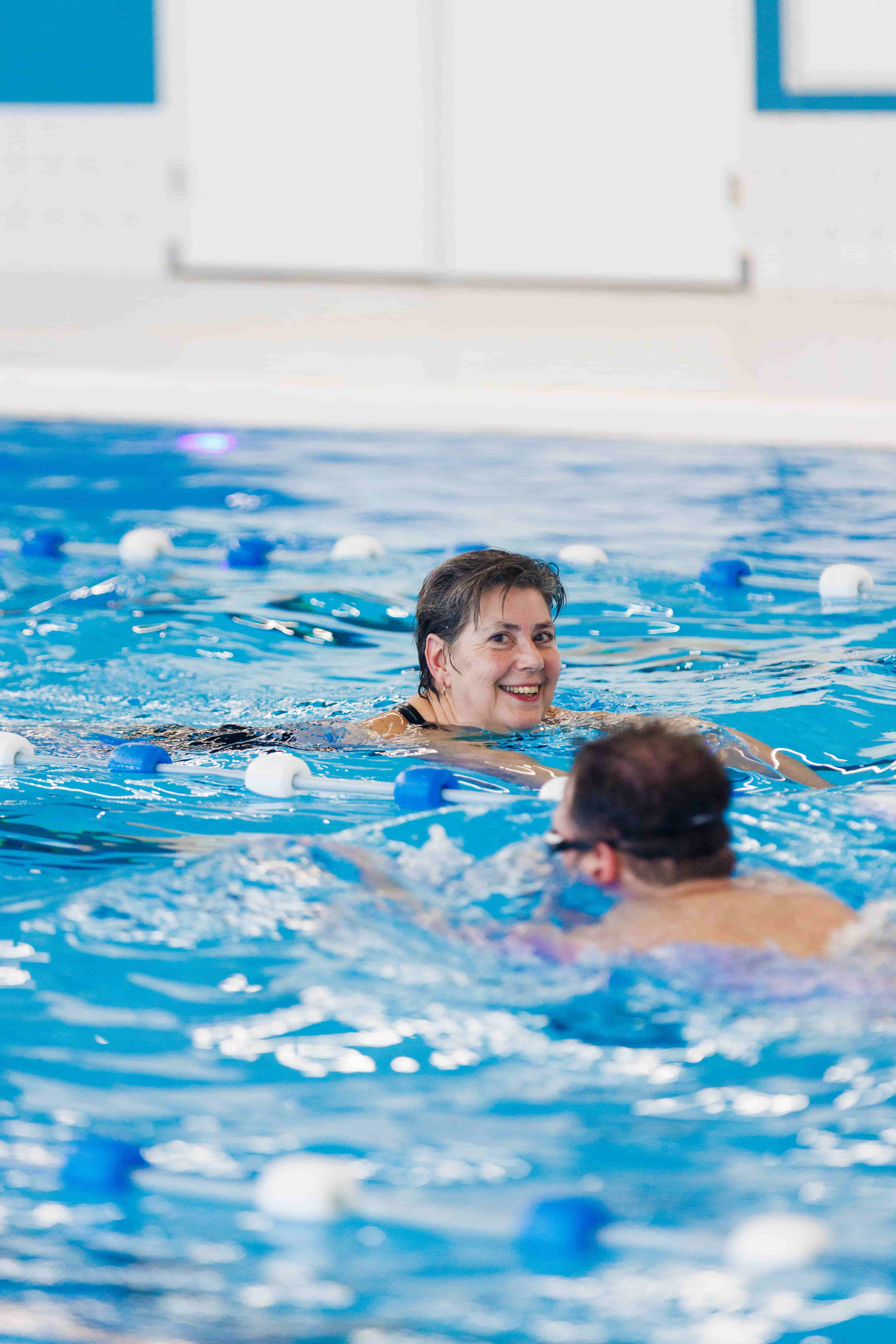 Tijdens het banenzwemmen bij Optisport kijkt een vrouw, terwijl ze de schoolslag zwemt, lachend naar de camera. 
