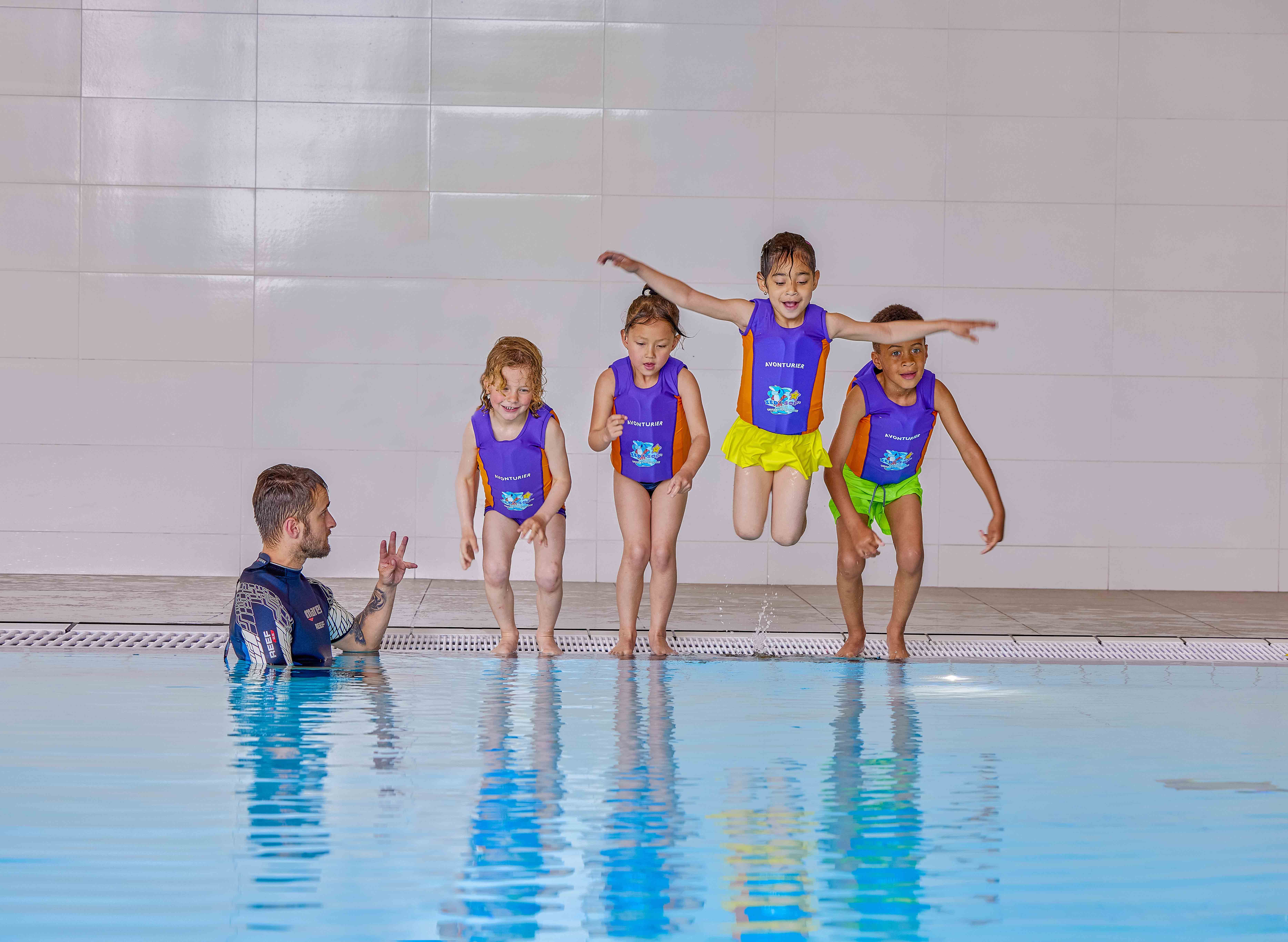 Kinderen springen met vestje in het diepe water, de instructeur van Optisport geeft uileg en kijkt goedkeurend toe. 