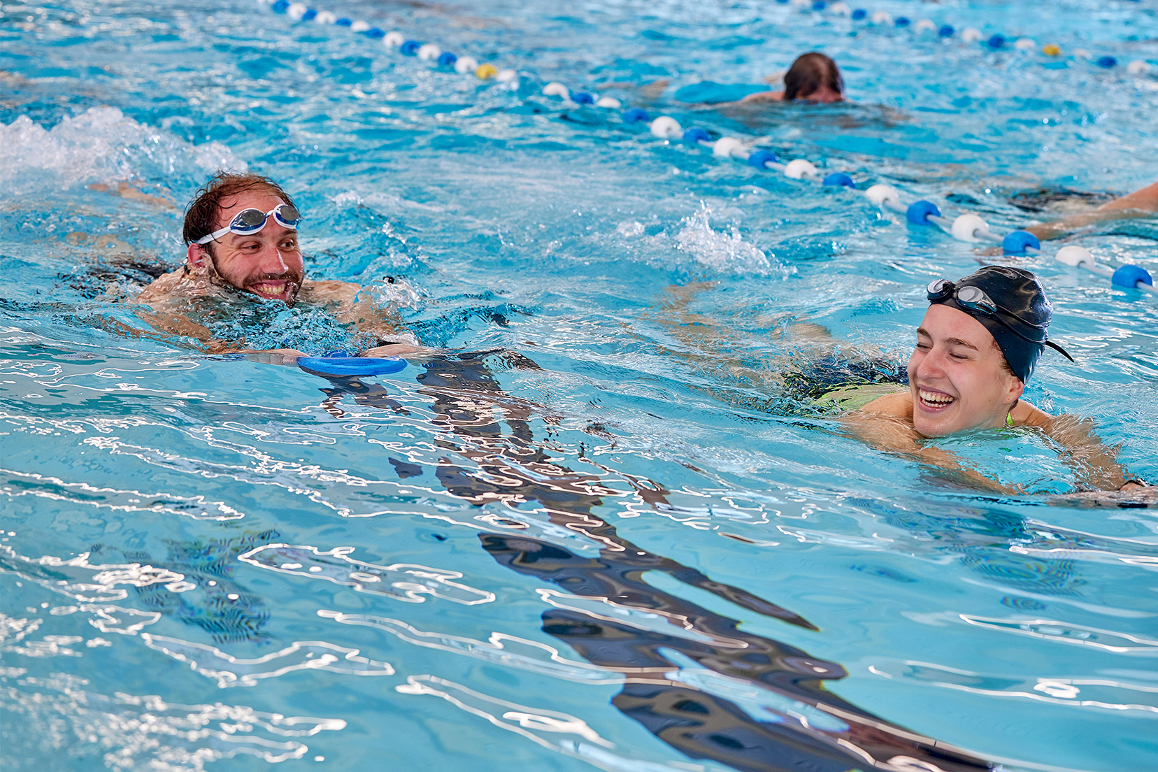 Twee ervaren zwemmers trainen gezamenlijk tijdens de les sportief banenzwemmen. Beide zwemmen benen met een plankje en hebben veel lol samen.