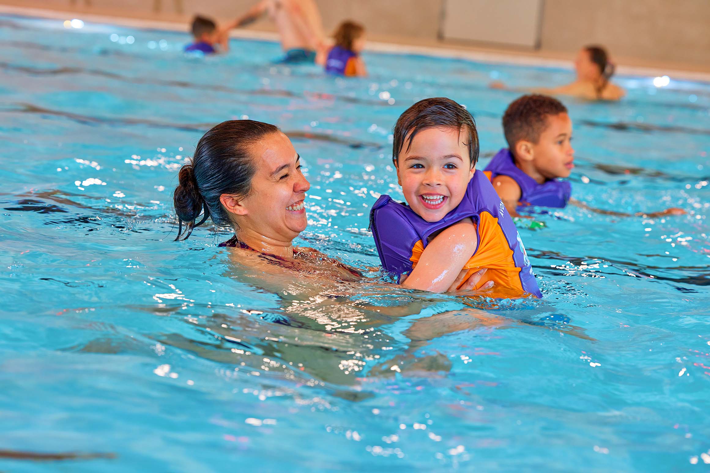 Een jongetje zwemt samen met zijn mama; hij draagt zijn Avonturier-zwemvest van de Zed en Sop zwemles en spettert er lekker op los. 