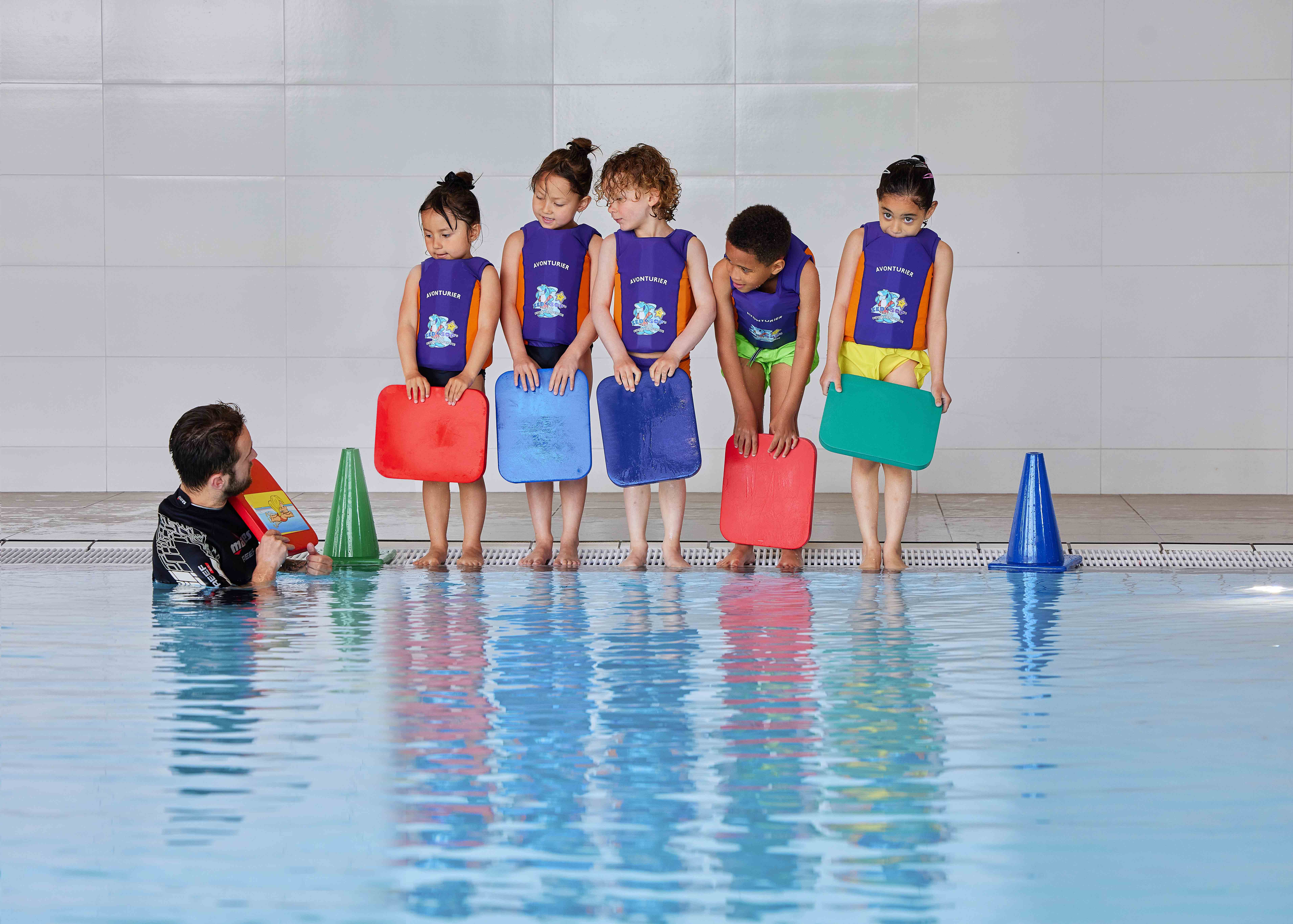 Vijf kindjes staan op de rand van het zwembad met hun Zed en Sop vestje aan, en kijken naar de instructeur die ze aanwijzingen geeft. 