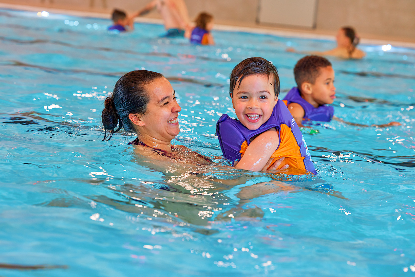 Een jongetje met een zwemvest van Optisport drijft in de armen van zijn moeder in het diepe water van het zwembad; samen maken ze veel plezier. 