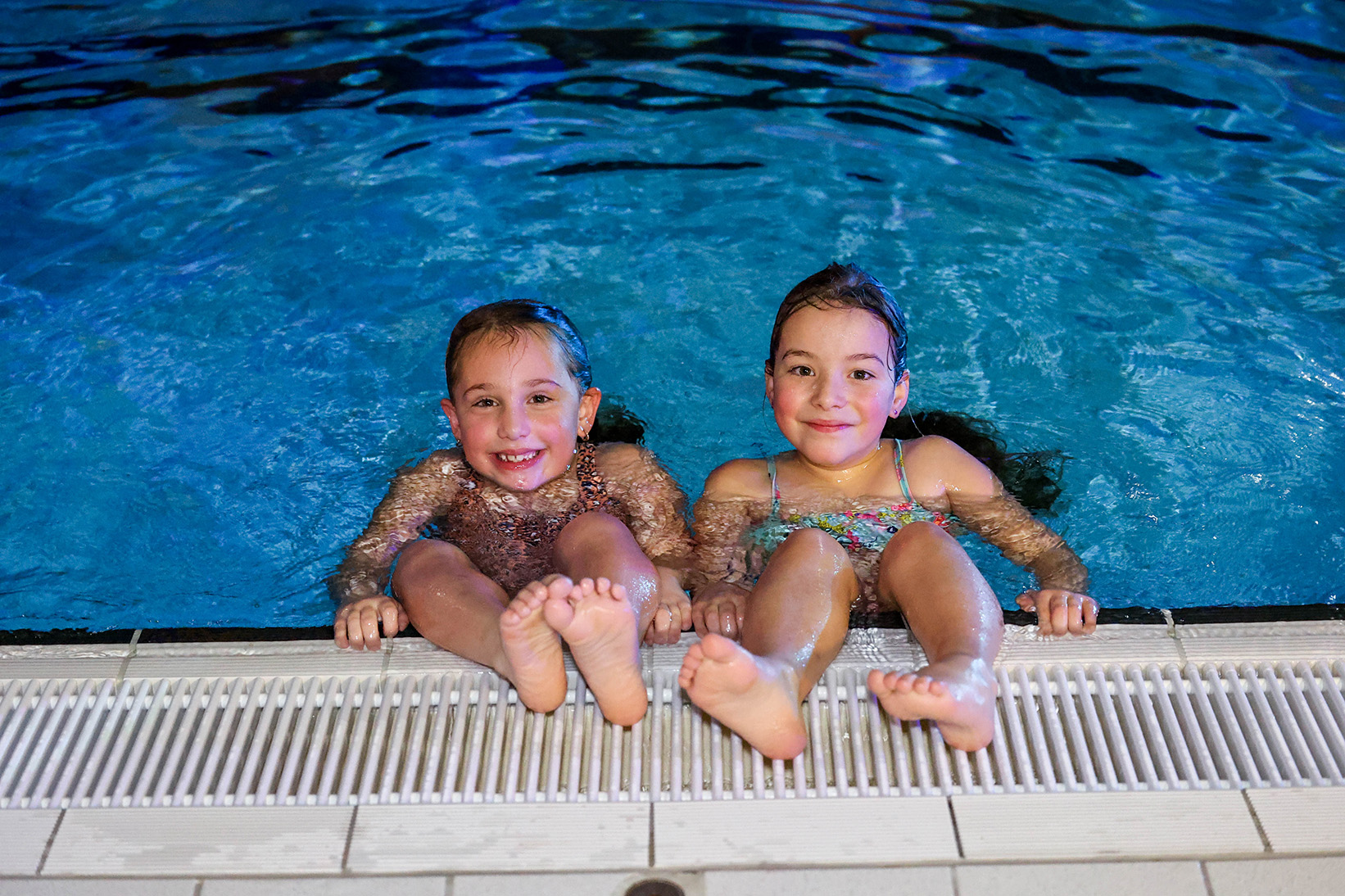 Twee meisjes hangen aan de rand van het zwembad van Optisport en kijken vrolijk in de camera: zij hebben een toptijd in het water! 