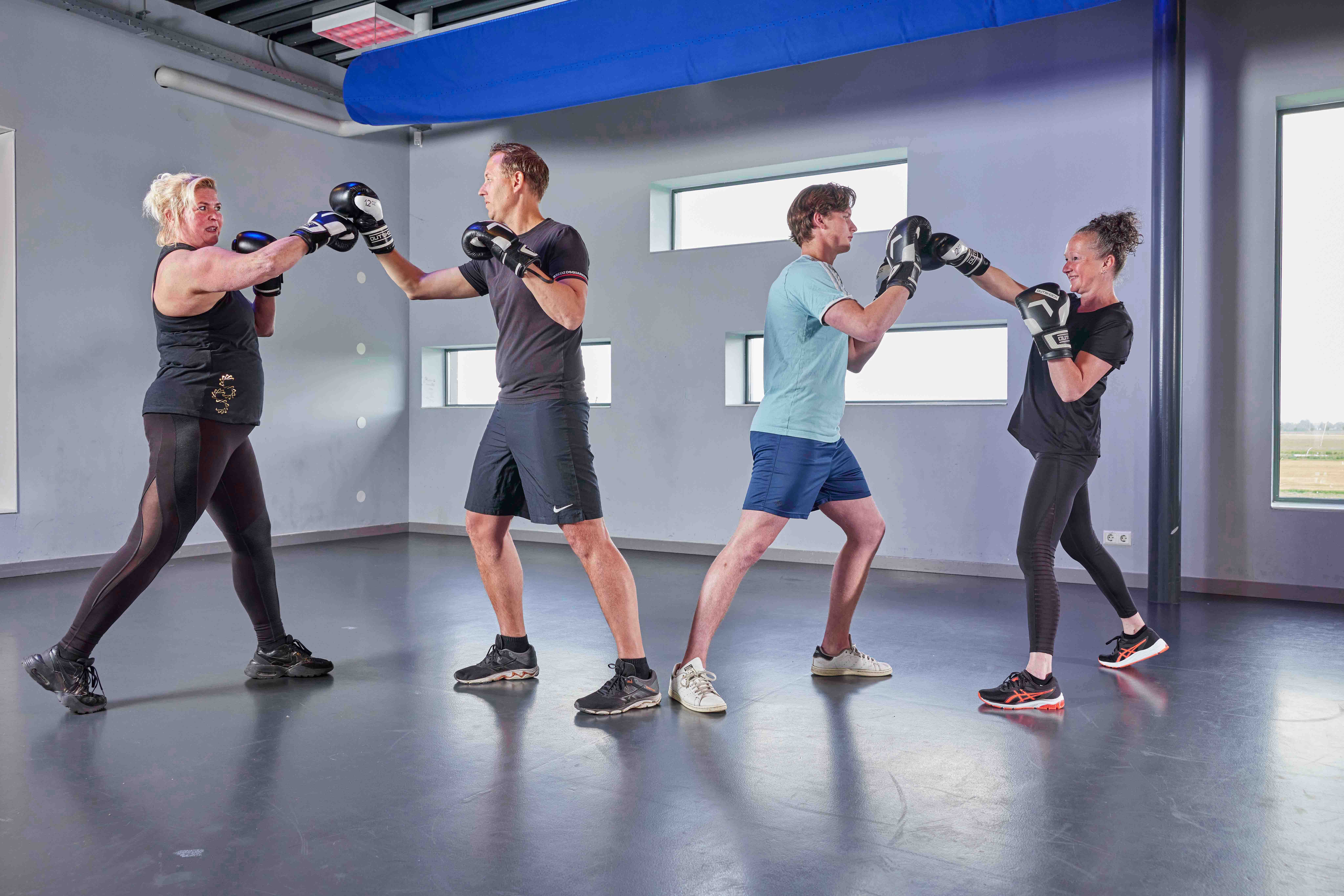 Twee mannen en twee vrouwen volgen een energieke boksles bij een Optisport healthclub, waarbij ze met elkaar sparren. 