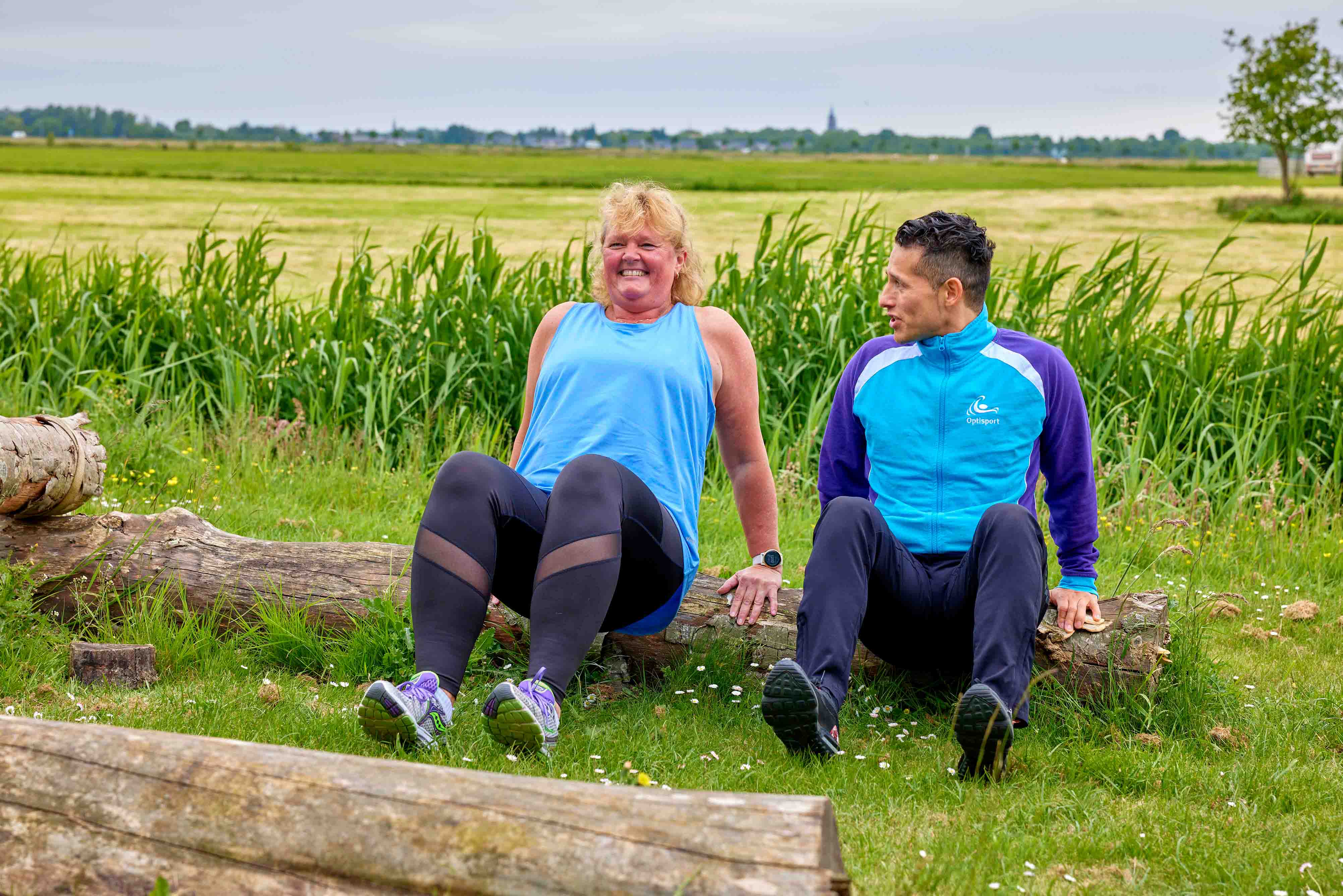 Een vrouw doet een krachtoefening tijdens de bootcamp training, terwijl ze wordt aangemoedigd door een enthousiaste instructeur van Optisport. 