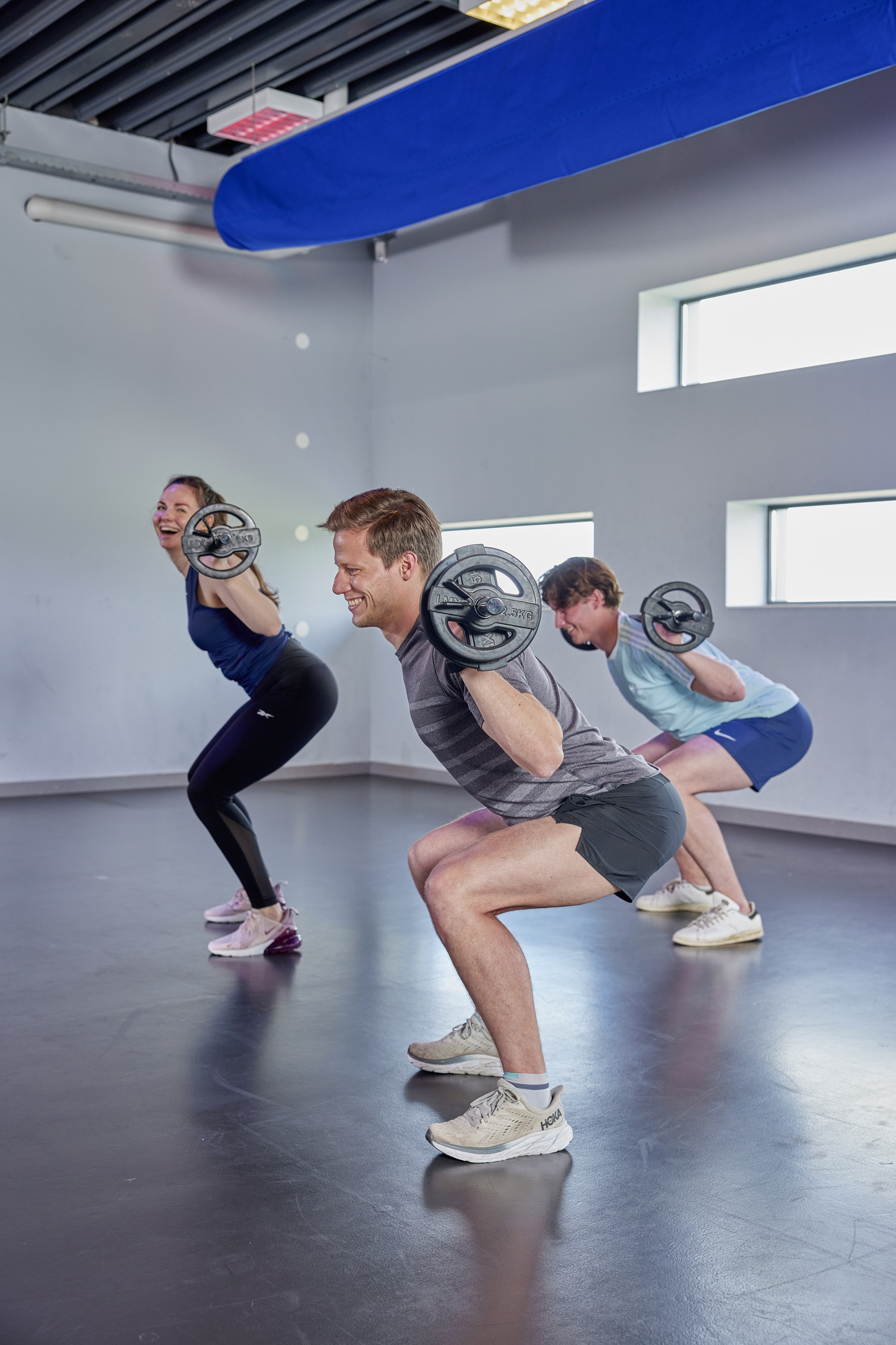 Drie sporters tillen ogenschijnlijk moeiteloos een barbell op hun nek terwijl ze kniebuigingen doen, tijdens een BootyFit les bij Optisport. 