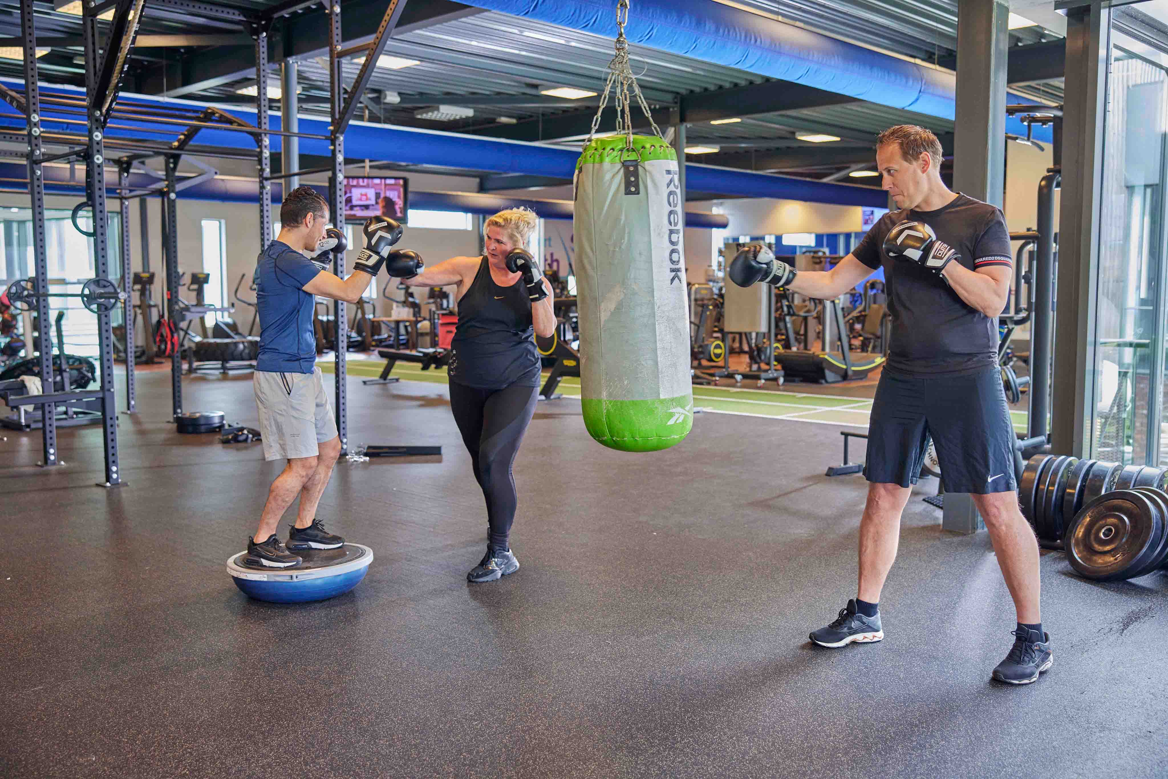 Twee sporters oefenen hun bokstechnieken; een samen met de Optisport instructeur en een op een grote boksbal tijdens een les Boxercise. 