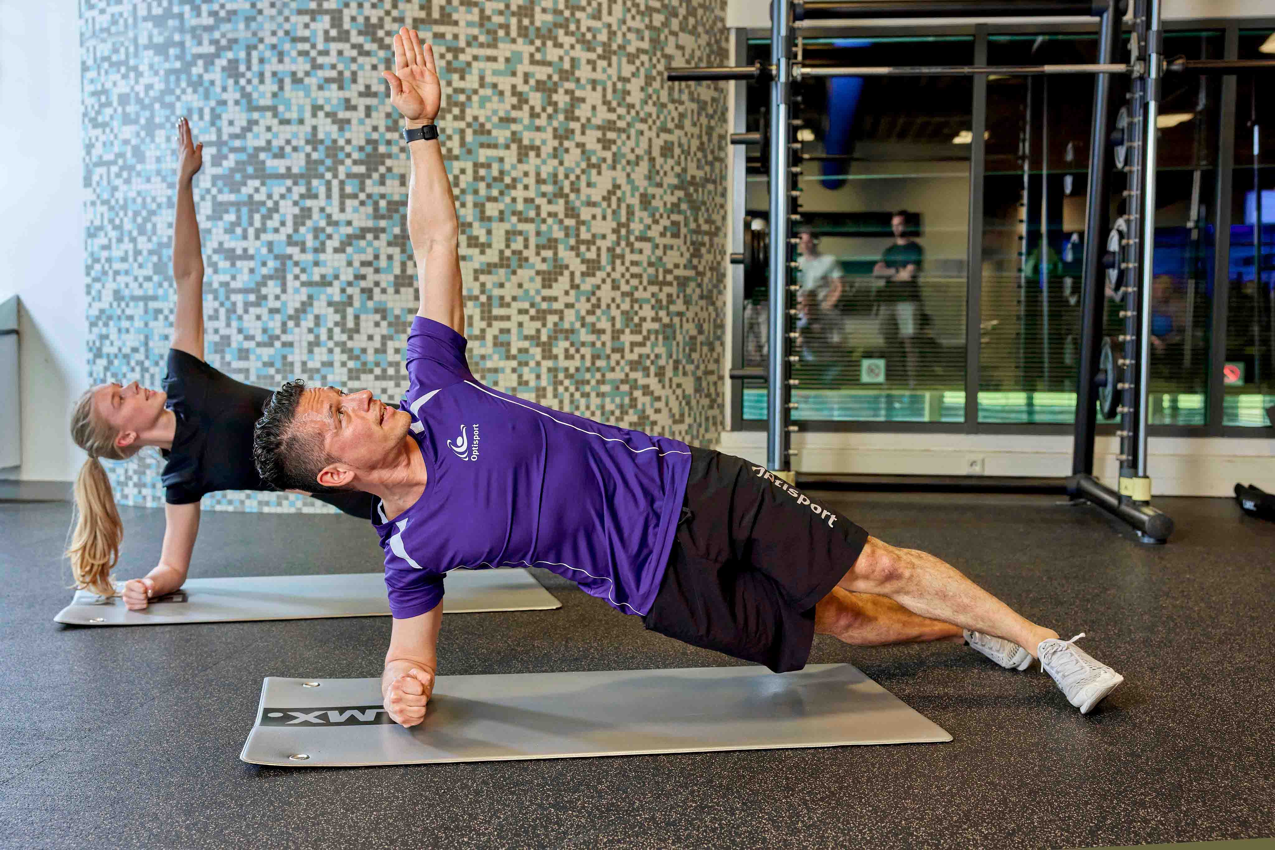 Een instructeur van Optisport doet samen met een andere sporter een zijwaartse strek- en krachtsoefening op een matje tijdens een les Buikspierkwartier bij Optisport.