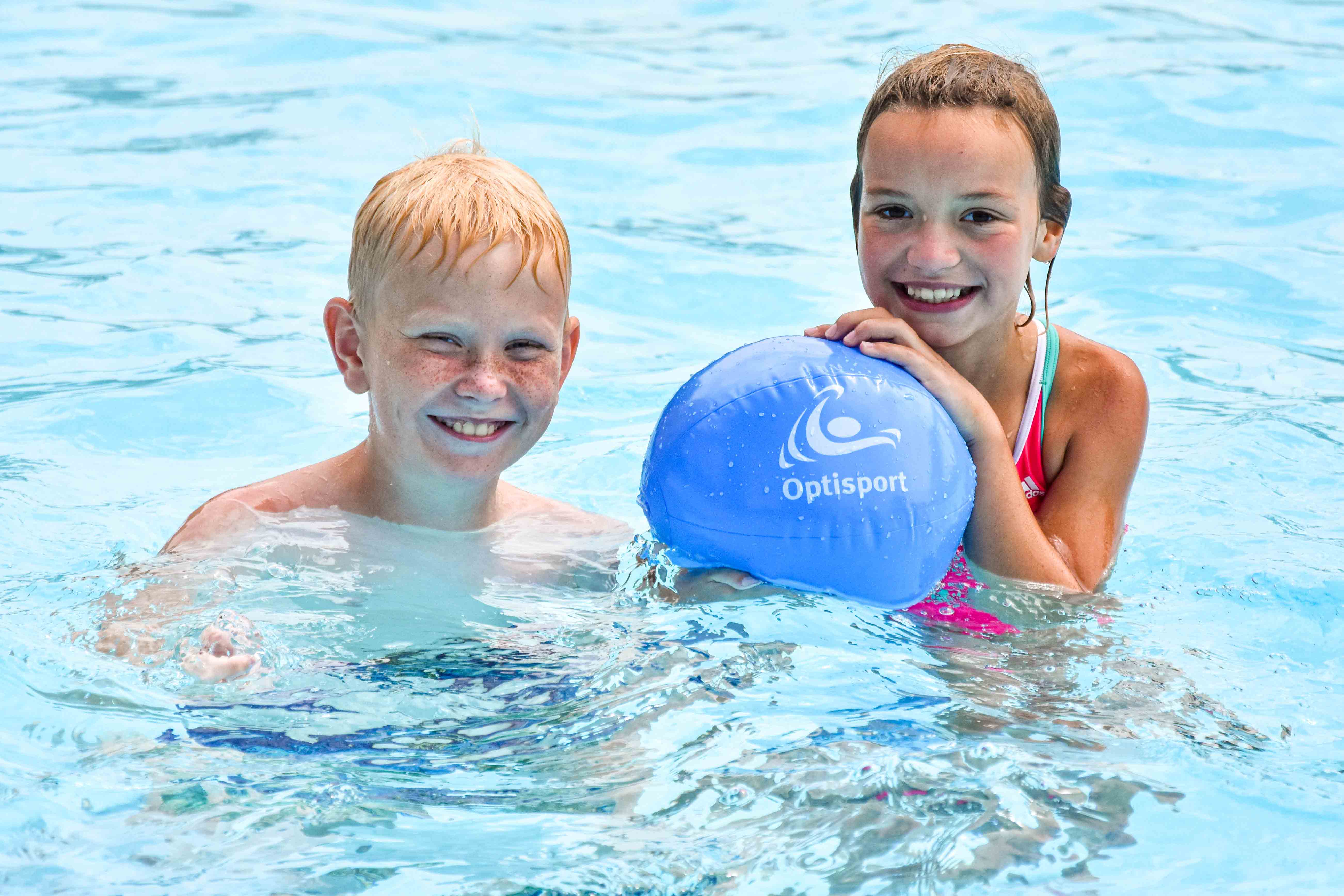 Een jongen en een meisje lachen naar de camera terwijl ze in een van de zwembaden van Optisport spelen met een strandbal. 