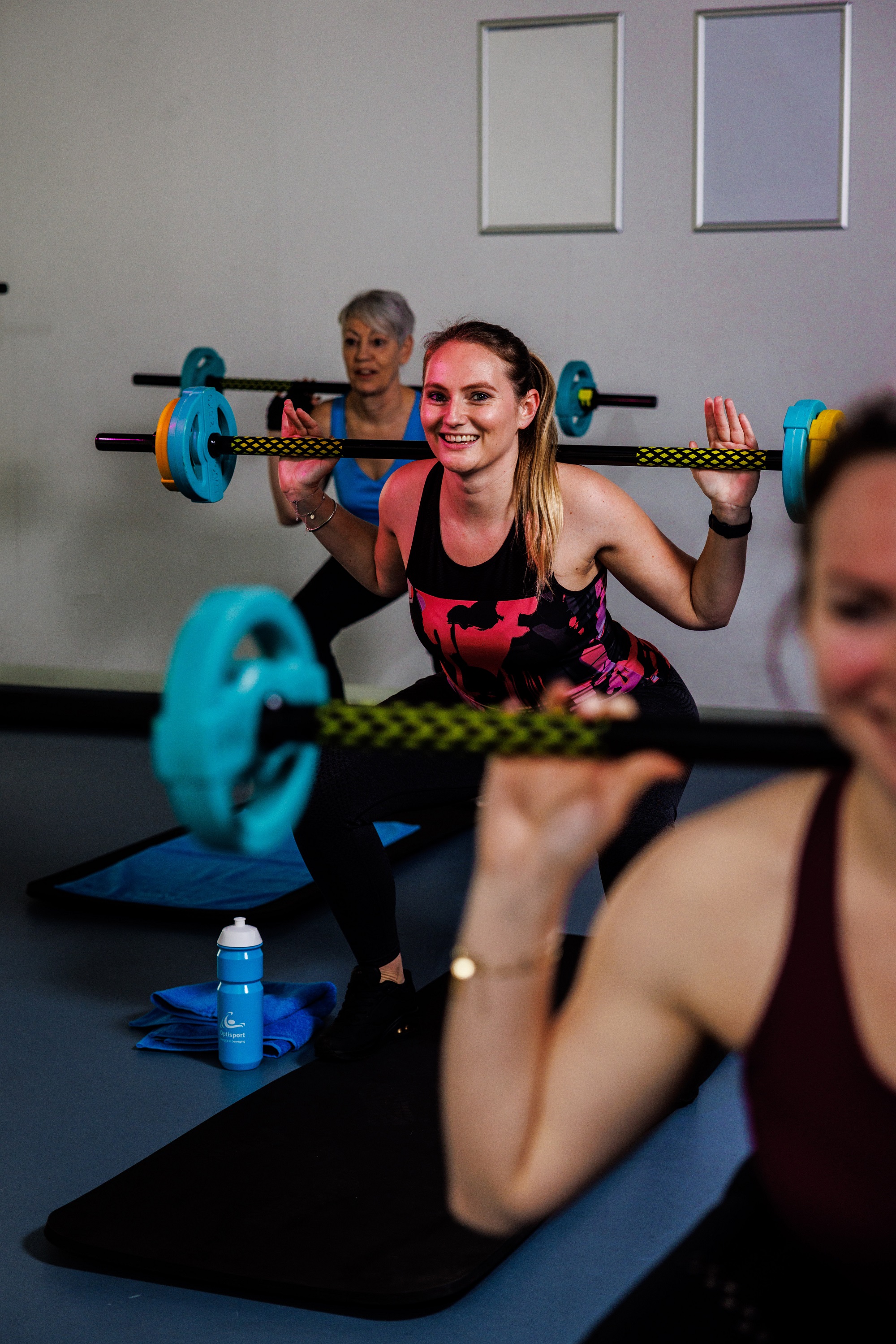 Drie vrouwen doen squats waarbij ze de barbell op hun nek omhoog tillen, tijdens de groepsles Club Power bij Optisport. 