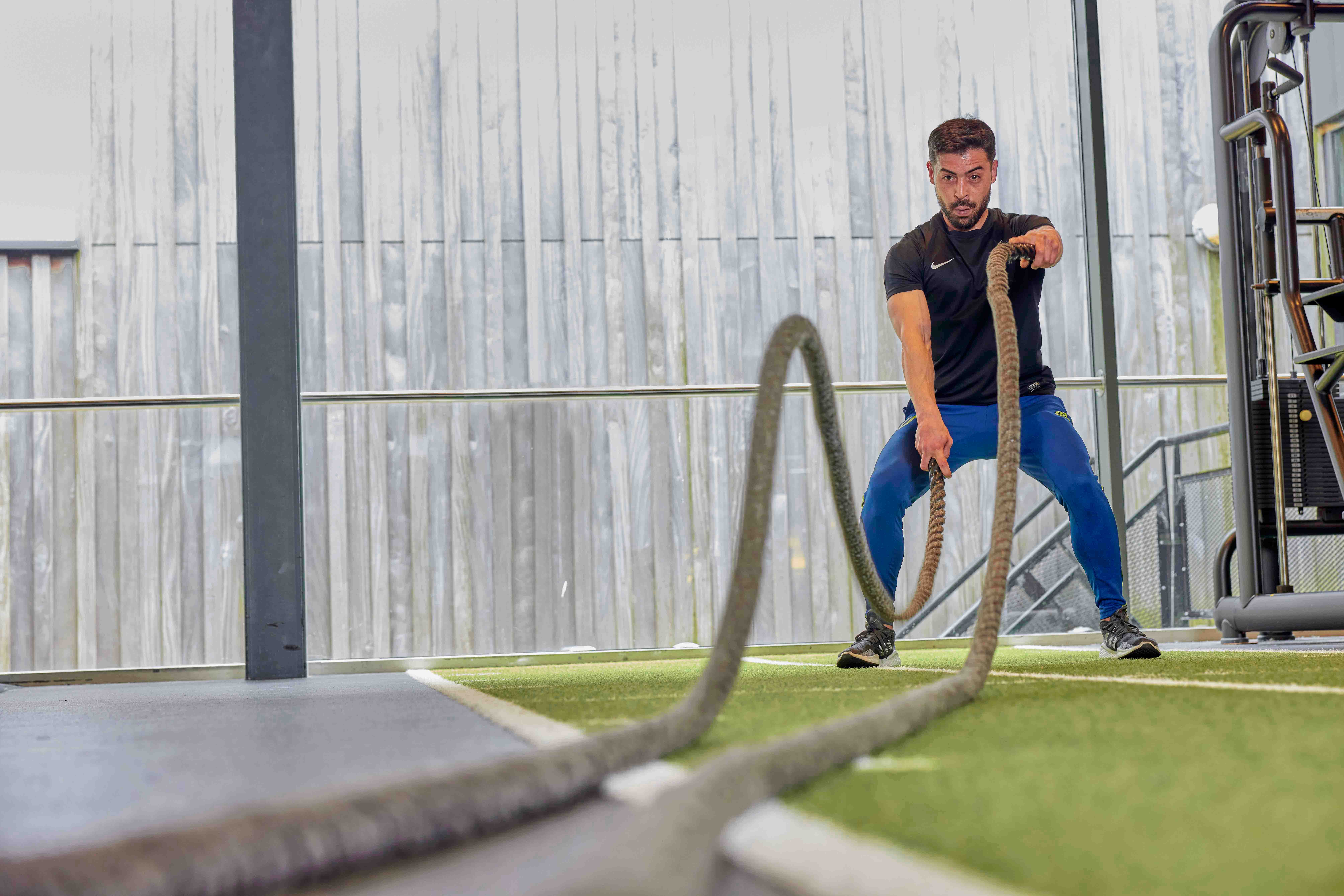 Een man is vol concentratie aan het trainen in een healthclub van Optisport; door zware touwen op en neer te bewegen, traint hij diverse spiergroepen. 
