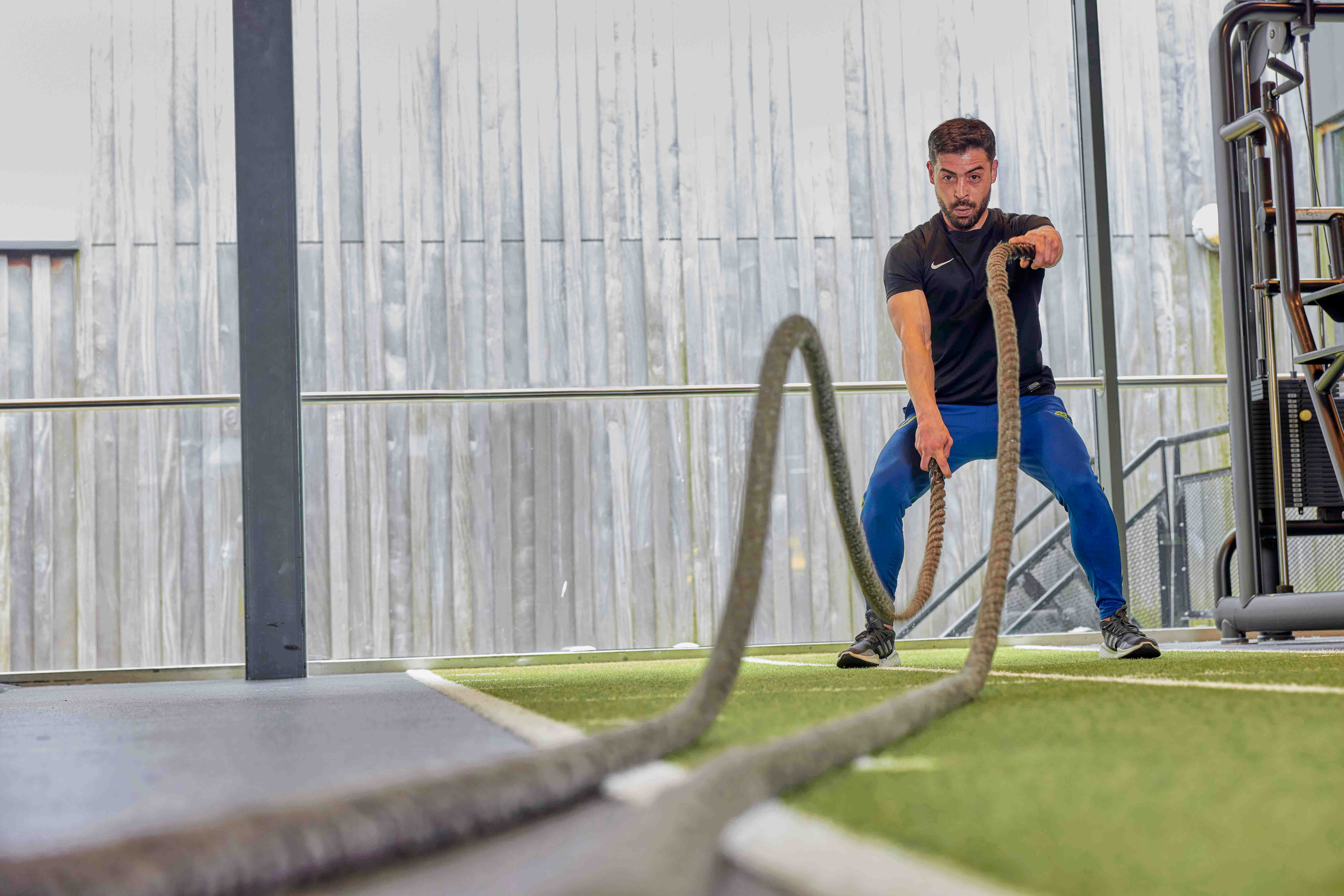 Een man is vol concentratie aan het trainen in een healthclub van Optisport; door zware touwen op en neer te bewegen, traint hij diverse spiergroepen.