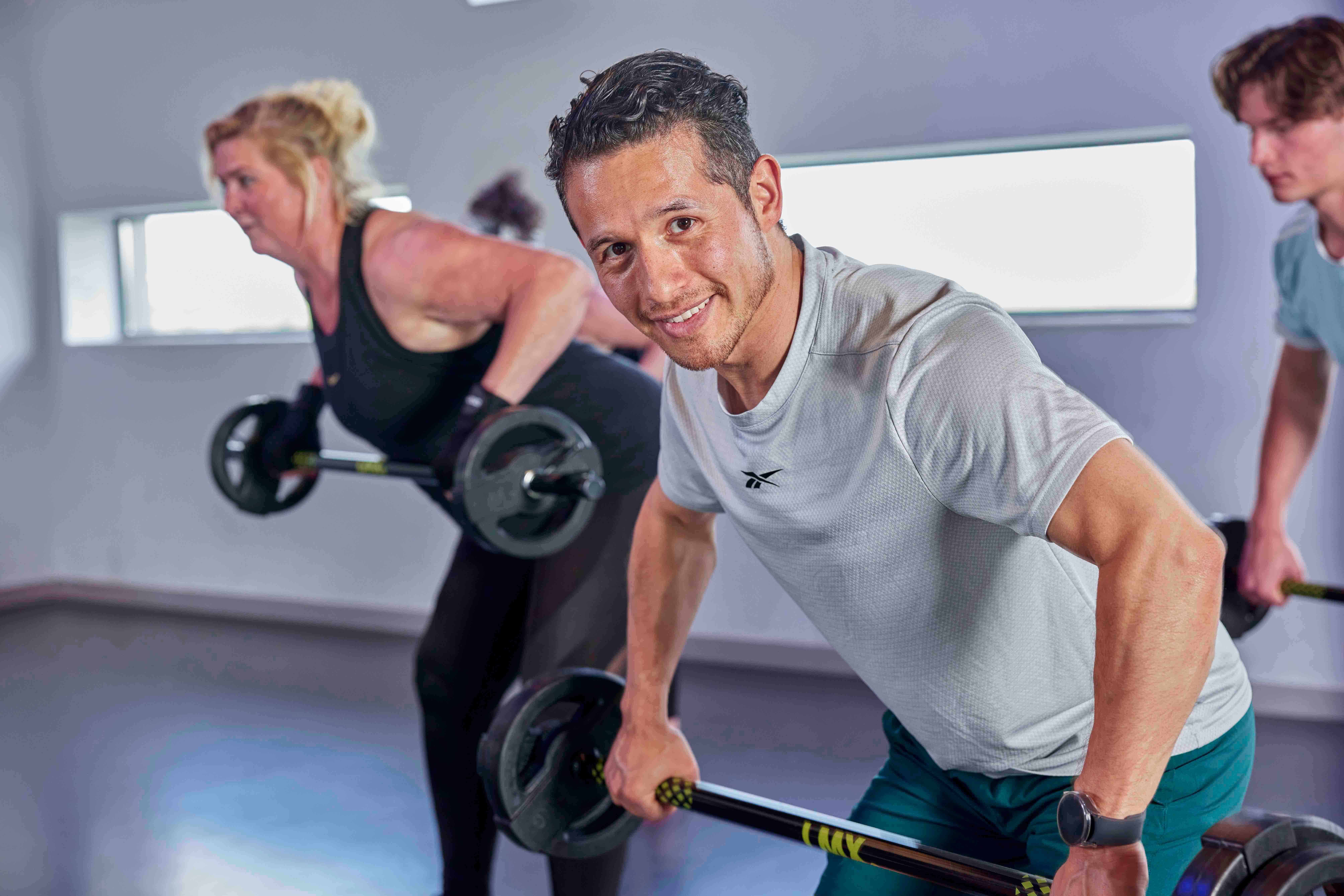 Een sporter kijkt lachend in de camera terwijl hij halters lift tijdens een van de groepslessen in een Optisport healthclub.