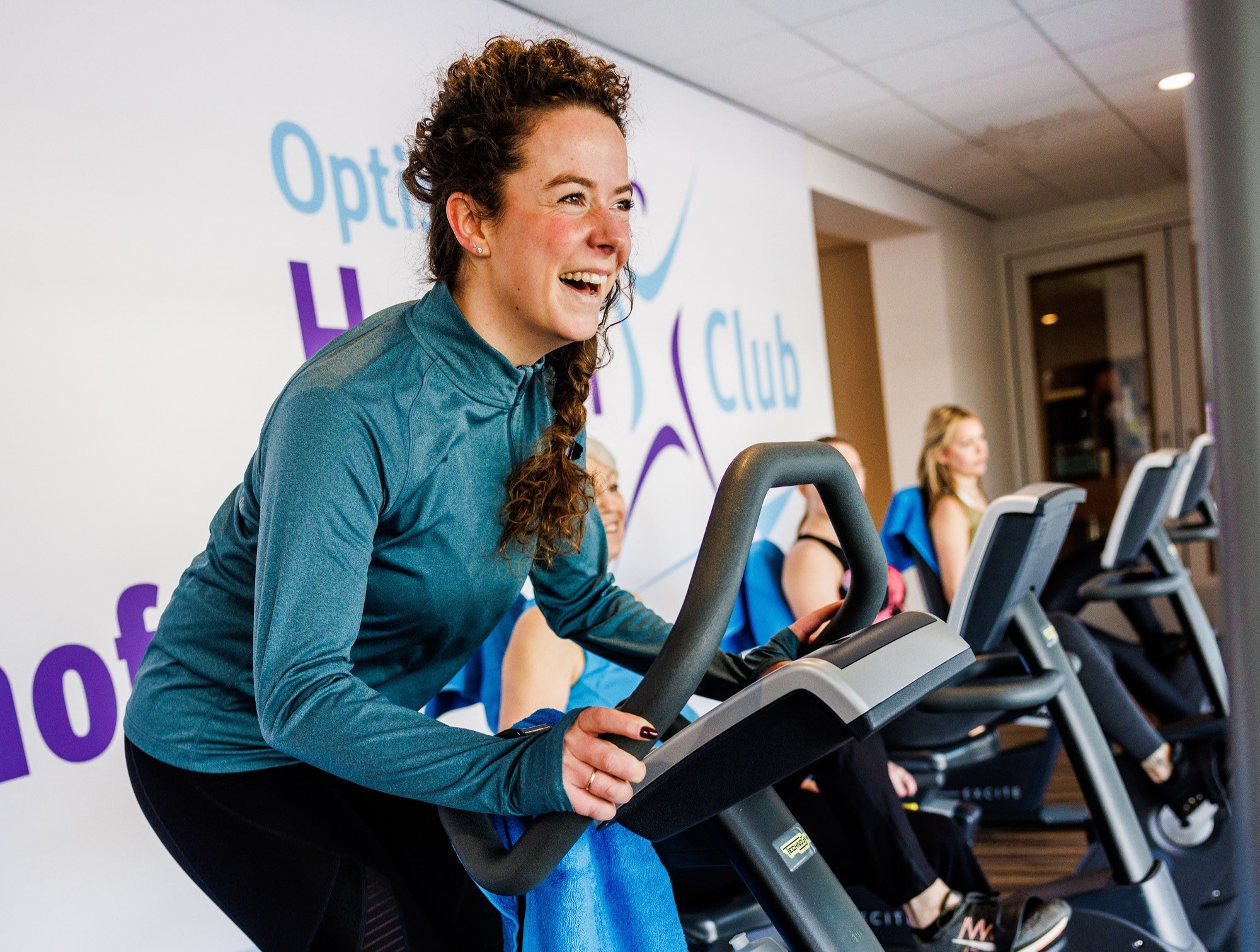 Een vrouw fietst met zichtbaar plezier op de indoor cycling apparaten in een van de healthclubs van Optisport. 