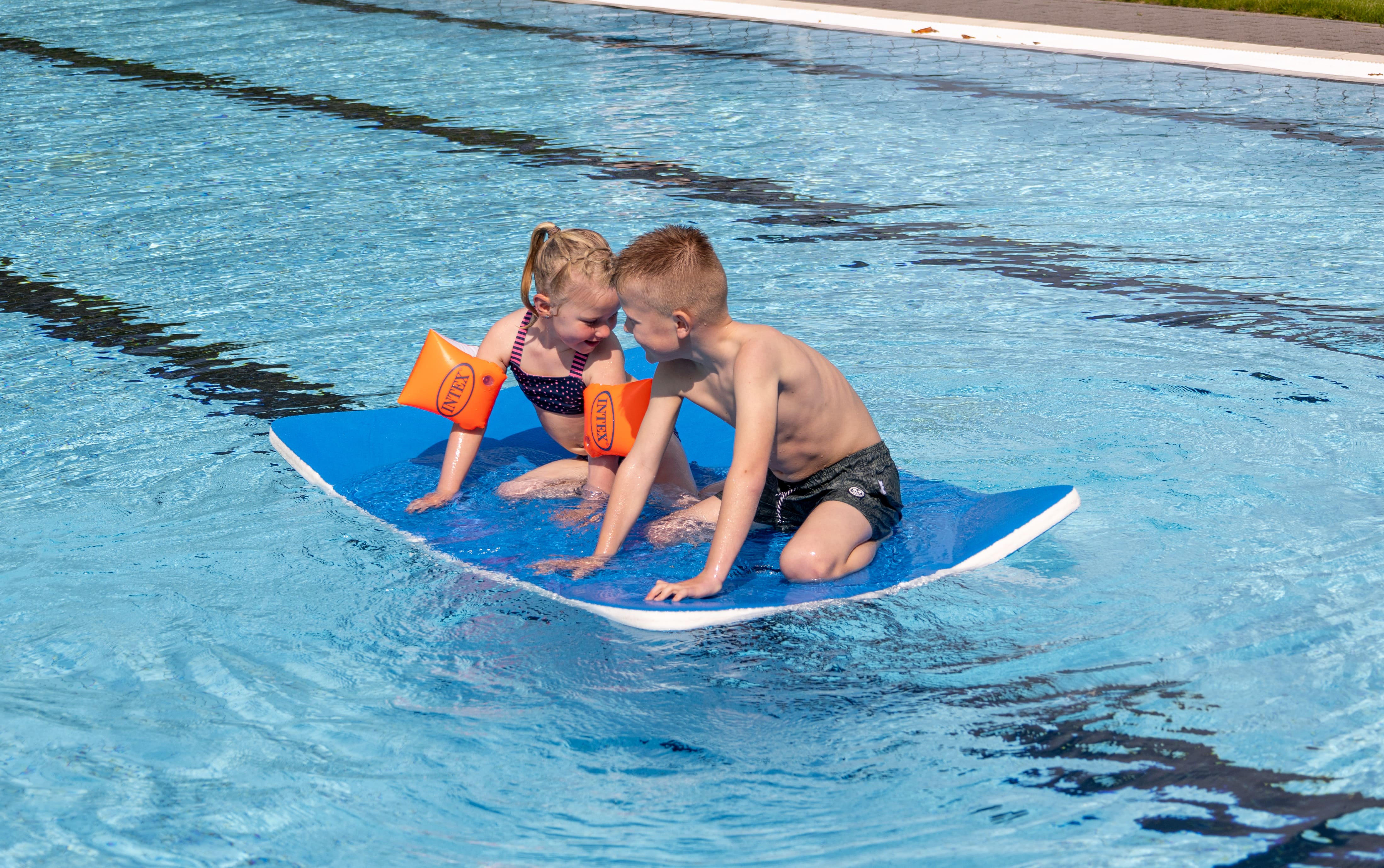 Twee kinderen zitten samen op een drijfmat in het buitenbad van Optisport; ze hebben een binnenpretje samen en leunen met hun voorhoofden tegen elkaar. 