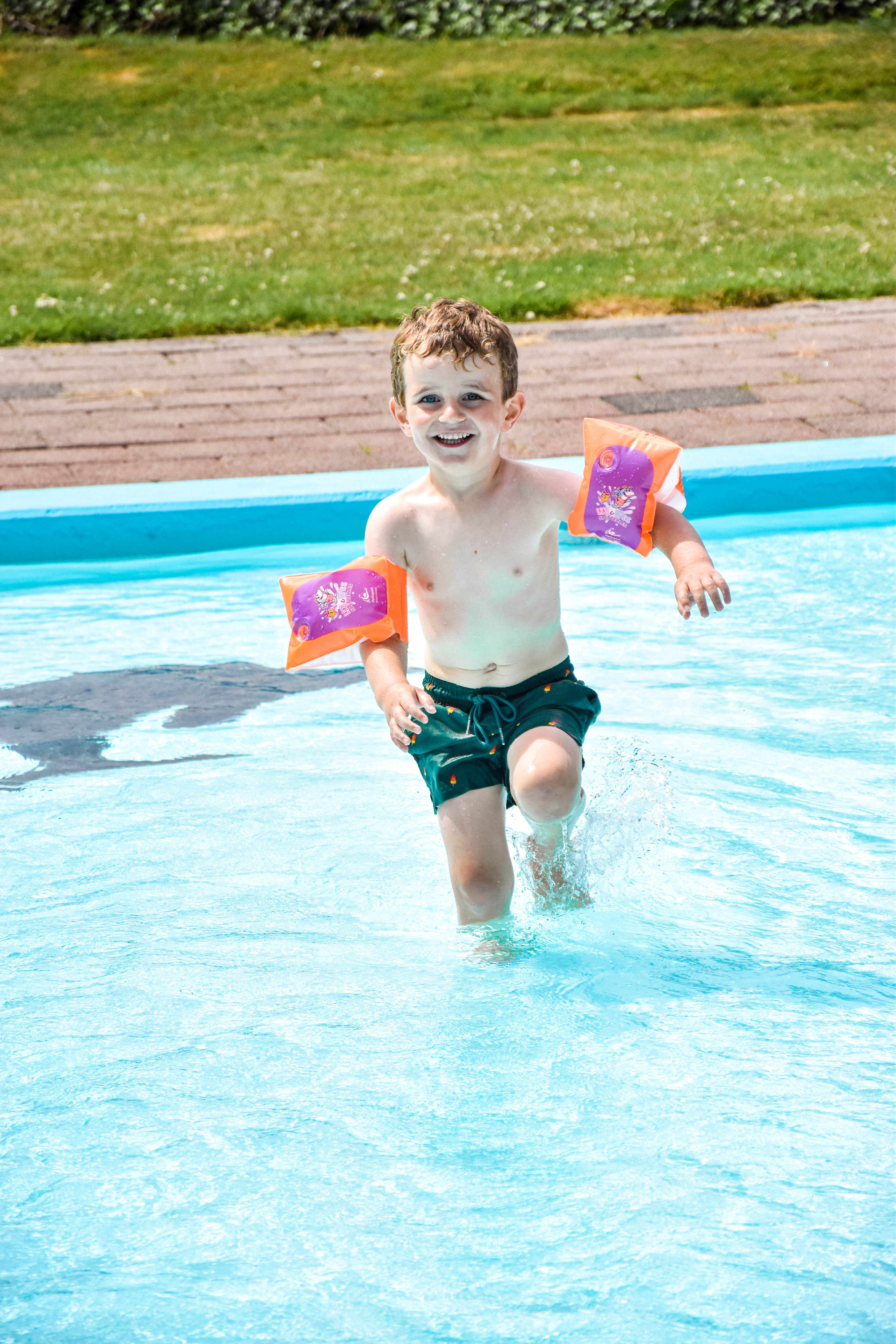 Een klein jongetje rent lachend door het ondiepe water tijdens het vrijzwemmen in het buitenbad in het buitenbad van Optisport, aan zijn armen prijken twee mooie Liz & Wes zwembandjes. 