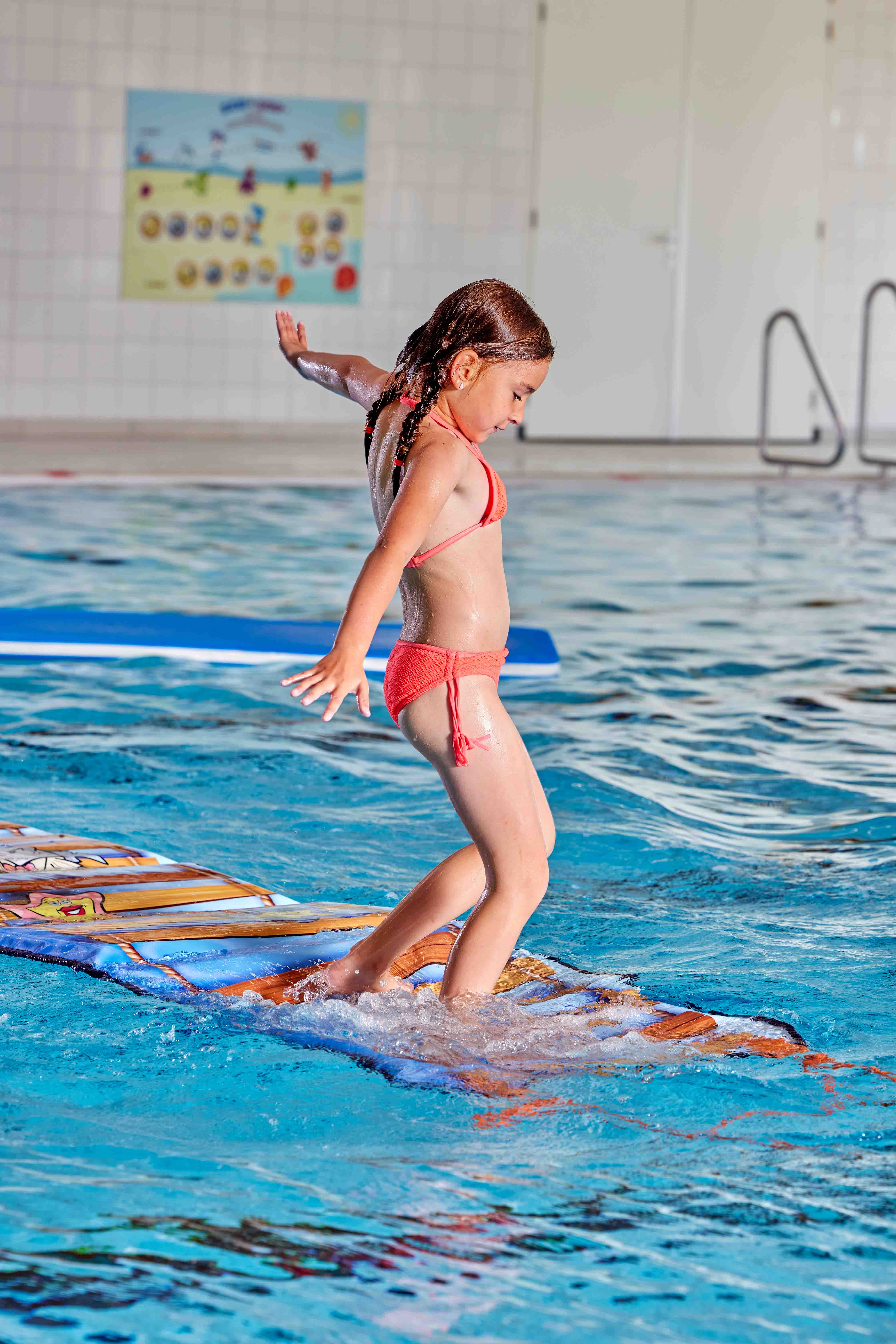 Een meisje probeert haar evenwicht te bewaren op een grote loopmat die over het zwembad is gelegd, tijdens het Vrijzwemmen in een zwembad van Optisport. 