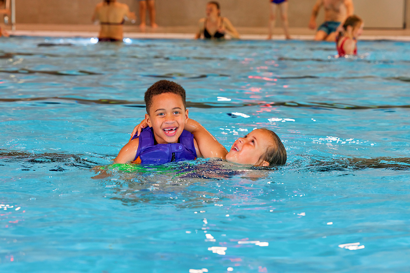 Twee kinderen spelen samen in het zwembad van Optisport tijdens het Vrijzwemmen; het jongetje draagt een zwemvest van Zed & Sop en het meisje zwemt zonder drijfmiddelen. 
