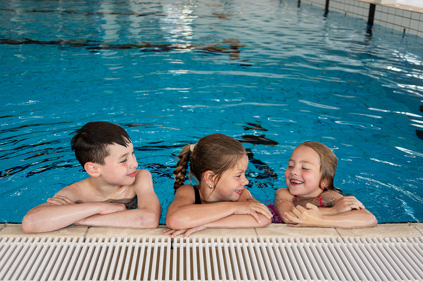 Drie kinderen hangen met hun armen op de rand van het zwembad en lachen met elkaar om iets grappigs, tijdens het Vrijzwemmen in het zwembad van Optisport. 