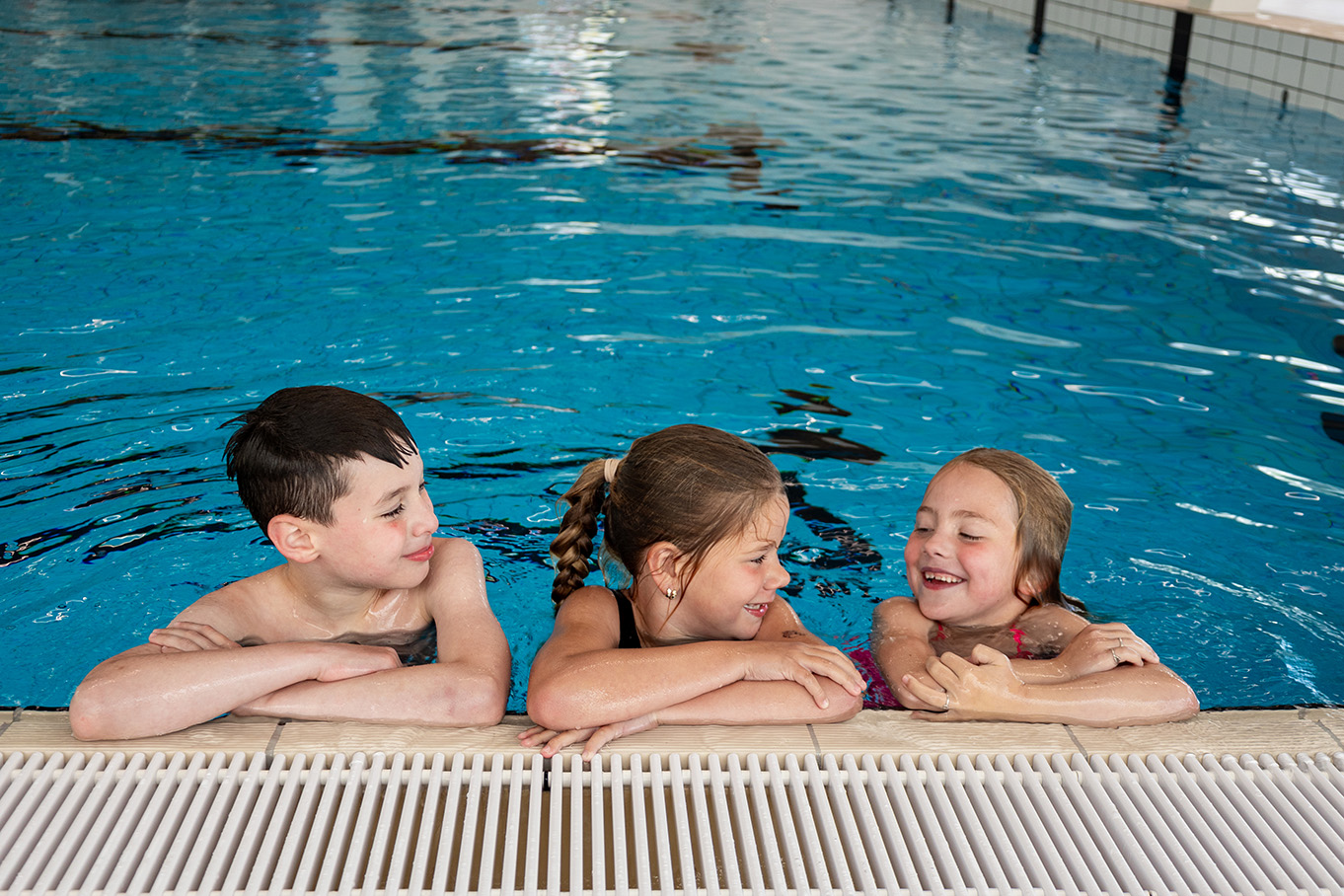 Drie kinderen hangen met hun armen op de rand van het zwembad en lachen met elkaar om iets grappigs, tijdens het Vrijzwemmen in het zwembad van Optisport.