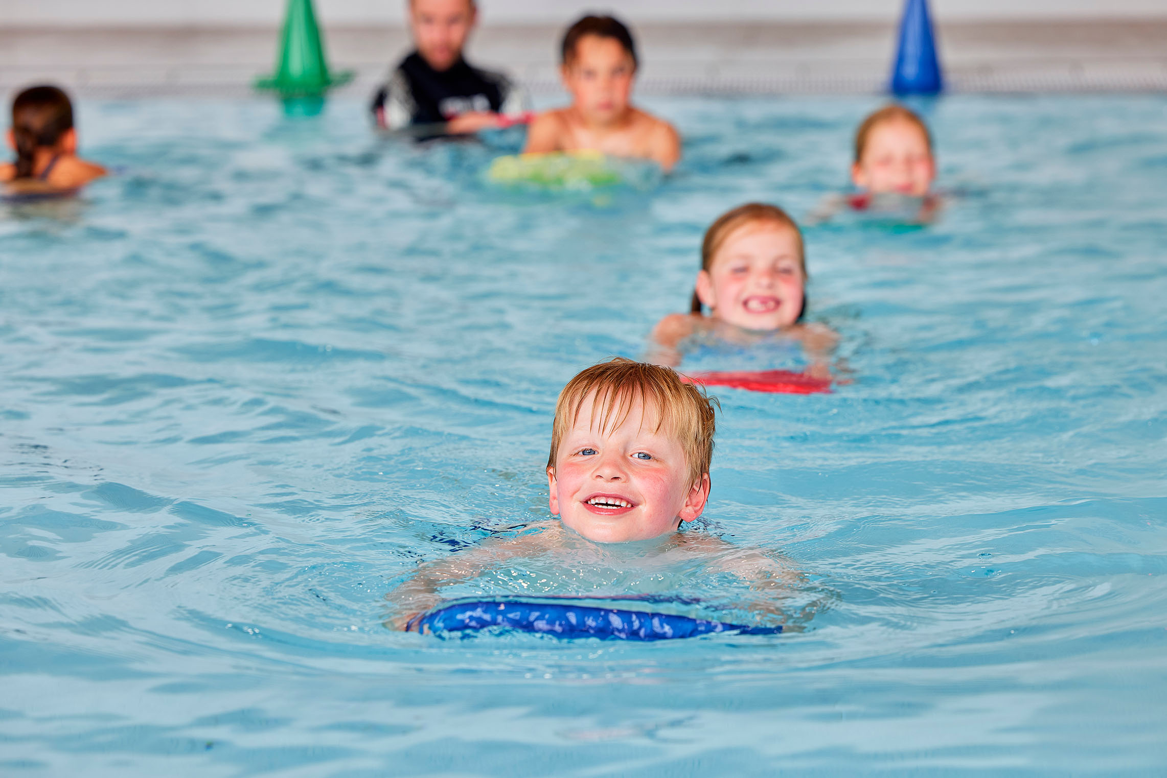 Kinderen zwemmen achter elkaar aan tijdens de zwemles van Optisport: in het diepe water, terwijl ze een drijfmiddel vasthouden maken ze plezier. 